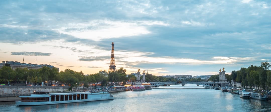 A twilight view of the Eiffel Tower from the Seine river, with tour boats, riverside quays, and bridges visible along the water. - Photo by Armands Brants on Unsplash