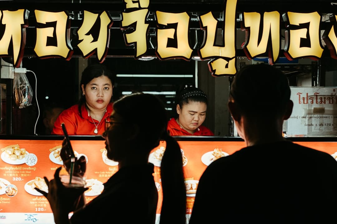 A street food vendor stall with two workers in red uniforms behind an orange counter displaying food items, with Japanese katakana signage and menu boards visible above. - Photo by Polina Kuzovkova on Unsplash