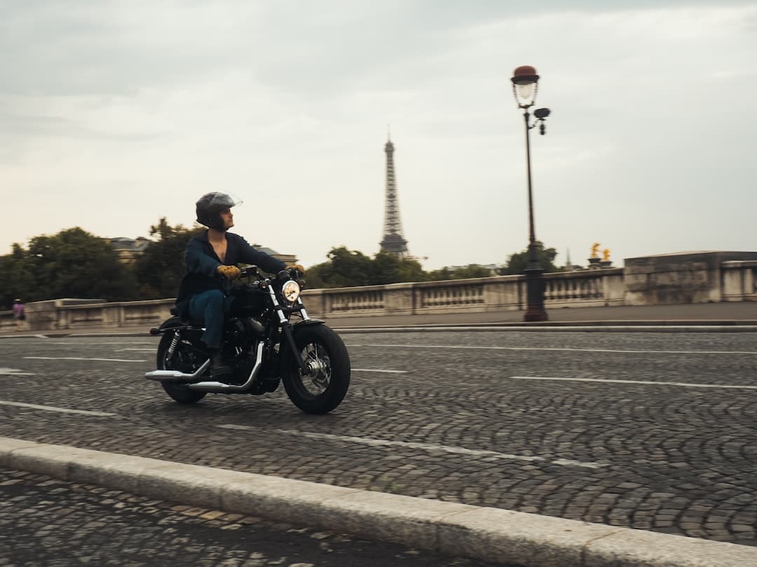 A person on a motorcycle rides across a cobblestone bridge in Paris with the Eiffel Tower visible in the background and a traditional street lamp to the right. - Photo by Lens by Benji on Unsplash
