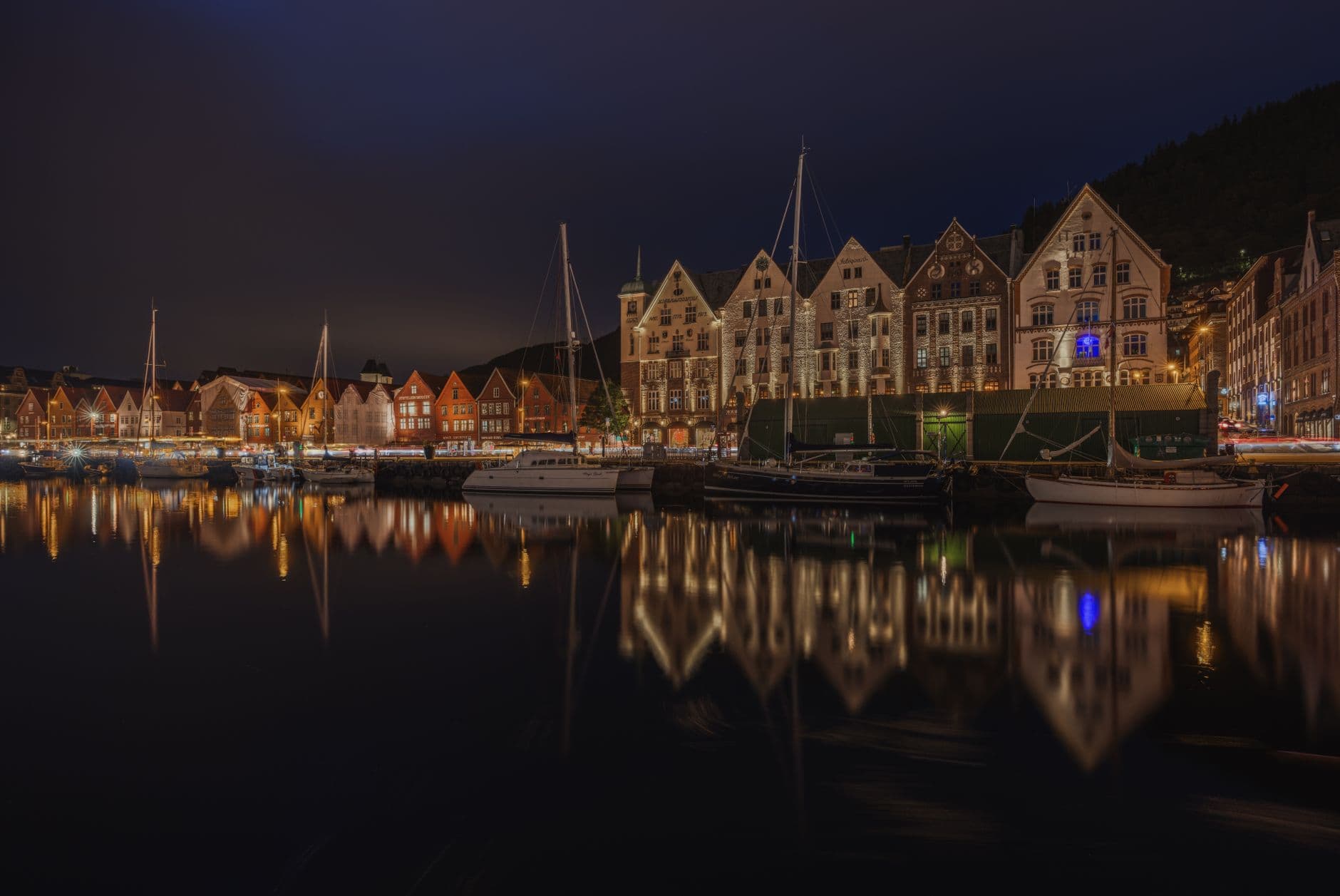 Night photograph of Bergen's historic Bryggen waterfront district illuminated against a twilight sky, with traditional Hanseatic merchant houses reflected in calm fjord waters and moored boats in the foreground. - Photo by Rino Adamo on Pexels