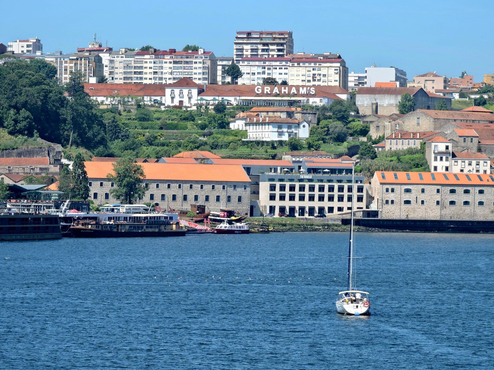 A waterfront view of Porto's Vila Nova de Gaia district across the Douro River, featuring the Graham's Port Lodge with its large signage, traditional stone warehouses with reconstructed roofs, modern residential buildings on the hillside above, and a sailboat moored in the foreground. - Photo by Rafael  Rodrigues on Pexels