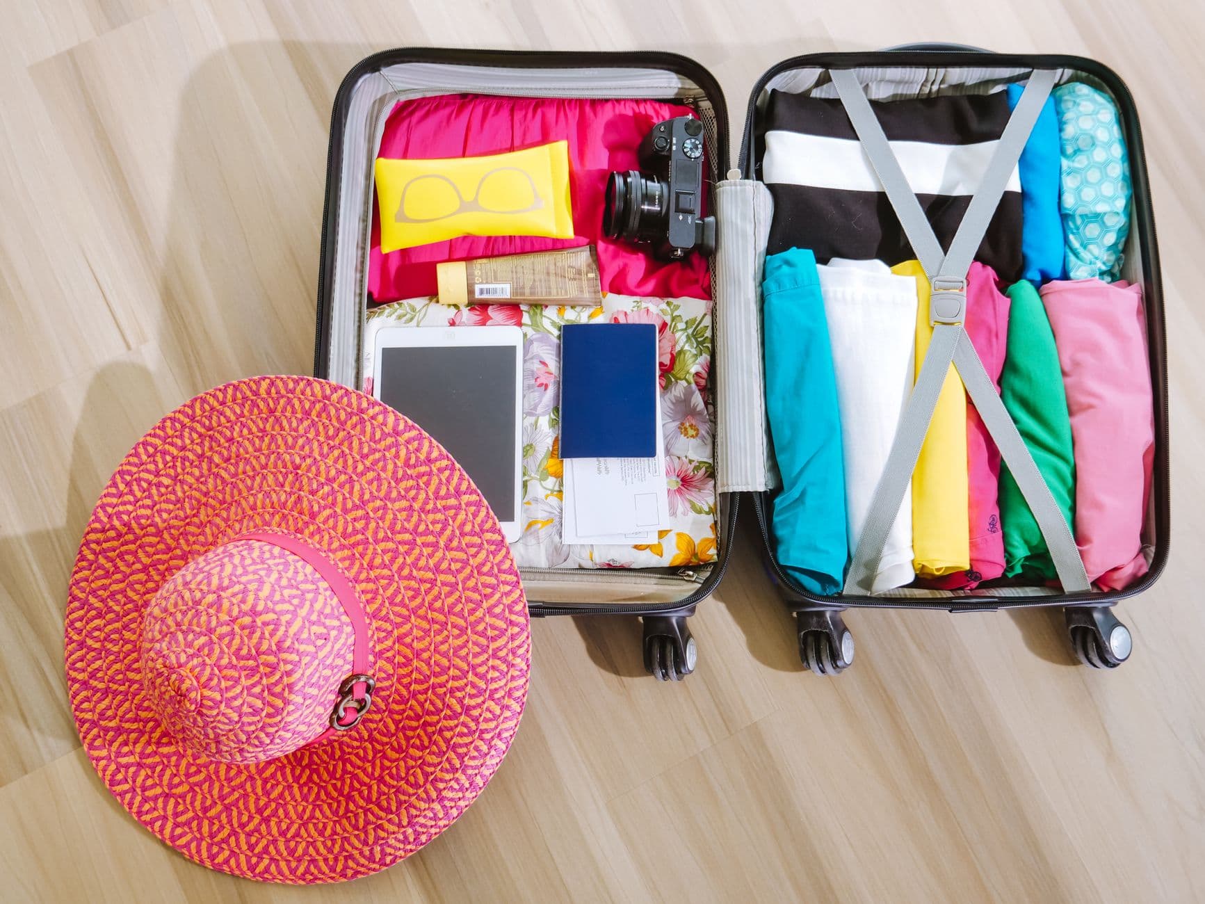 Overhead shot of two open suitcases packed with colorful clothing and travel accessories, with a pink sun hat beside them, photographed on a light wooden floor. - Photo by Kindel Media on Pexels