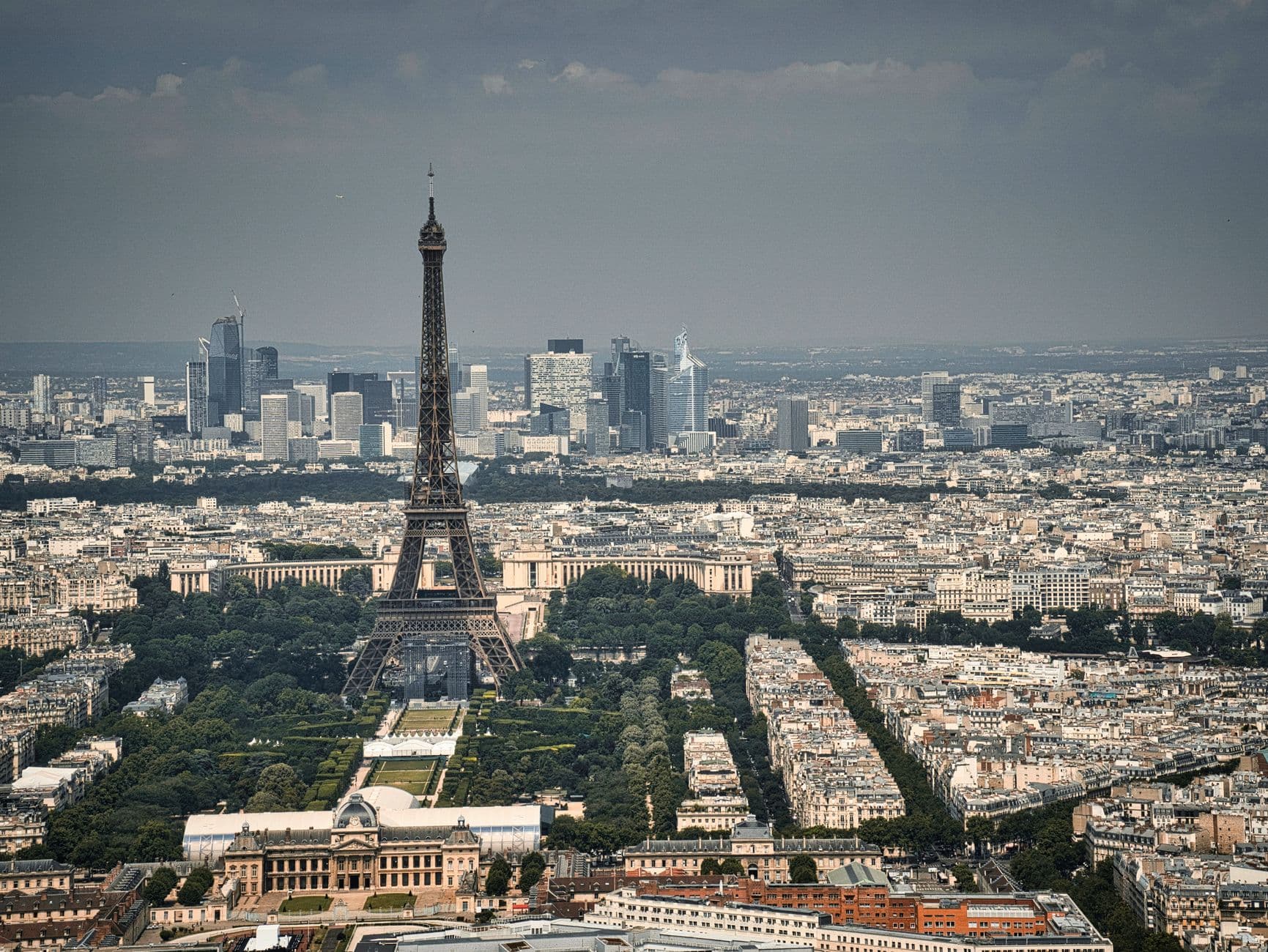 An aerial photograph of Paris taken from a high vantage point showing the Eiffel Tower dominating the center, with the Trocadéro in the immediate foreground, the Seine valley, and the broader Paris cityscape extending toward La Défense in the distance. - Photo by Travel with Lenses on Pexels