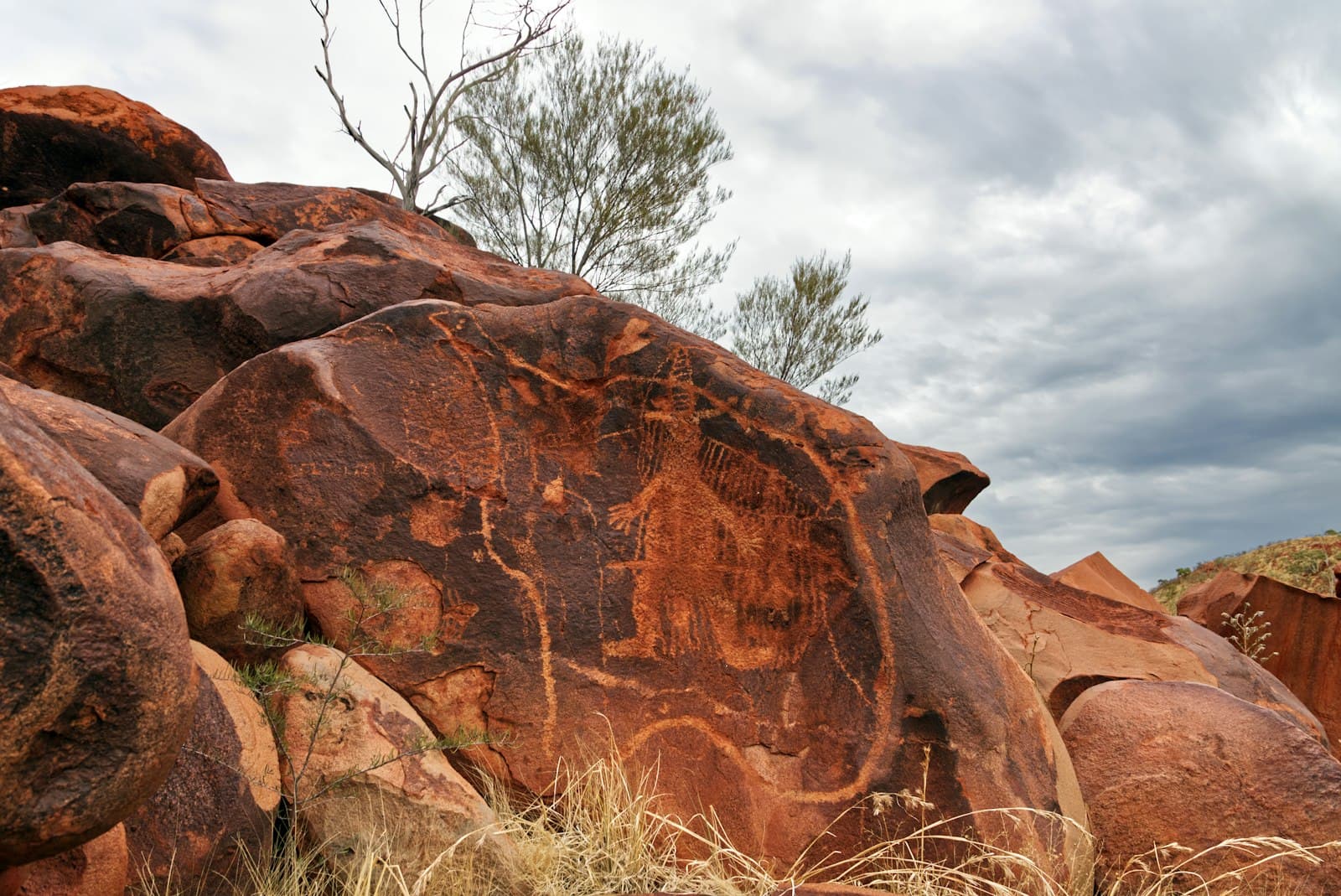 Ancient petroglyphs carved into red granite boulders on the Burrup Peninsula, showing human and animal figures with the characteristic patina and weathering of Aboriginal rock art sites. - Photo by René Riegal on Unsplash