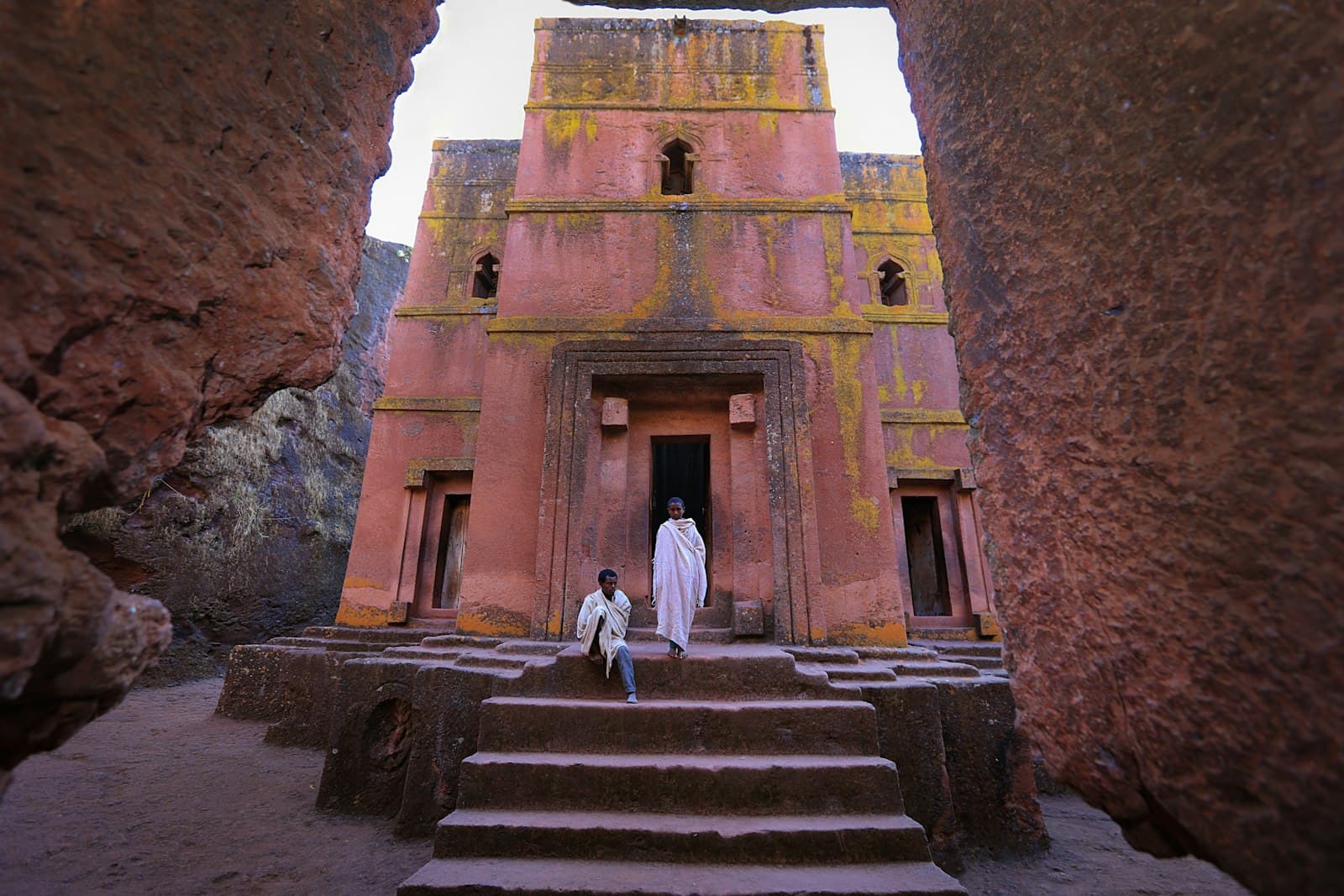 The Church of Saint George carved from red volcanic stone in Lalibela, Ethiopia, viewed from its sunken courtyard, with two figures in traditional white Ethiopian dress standing before its recessed façade. - Photo by mulugeta wolde on Unsplash