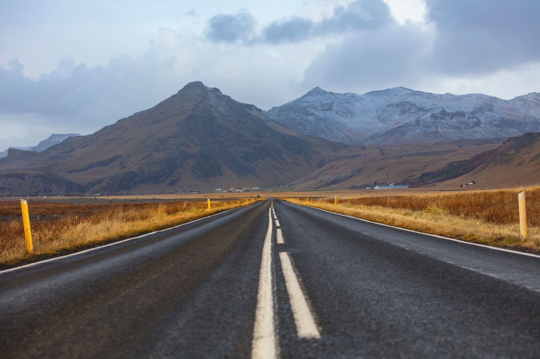 A straight asphalt road with white center line markings stretches toward distant snow-capped volcanic mountains across a barren, golden-brown Icelandic landscape flanked by yellow reflective posts. - Photo by Stephen Leonardi on Pexels