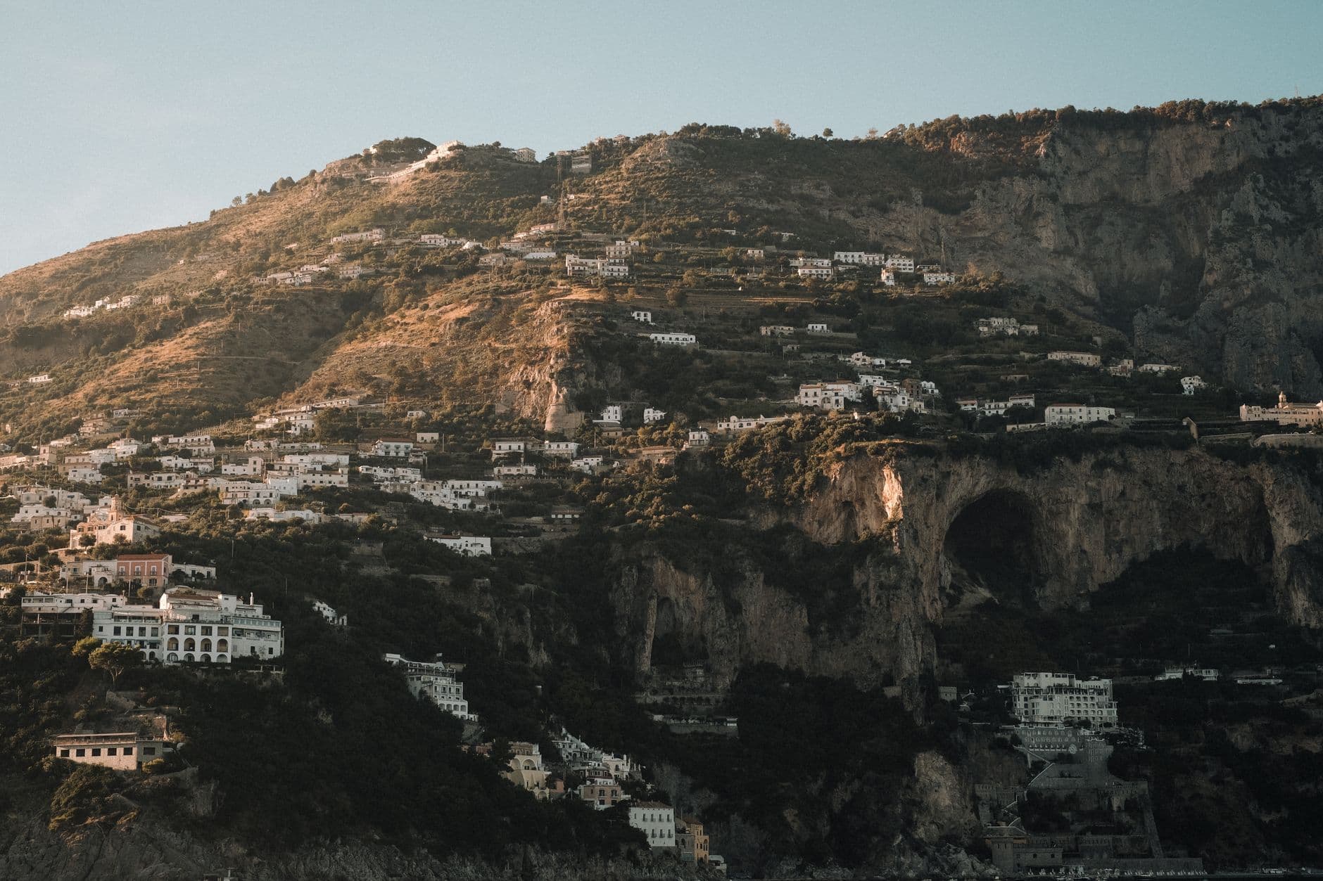A hillside village built into dramatic limestone cliffs with white and pastel-colored cubic buildings cascading down the slope, interspersed with Mediterranean vegetation. - Photo by Alina Chernii on Pexels