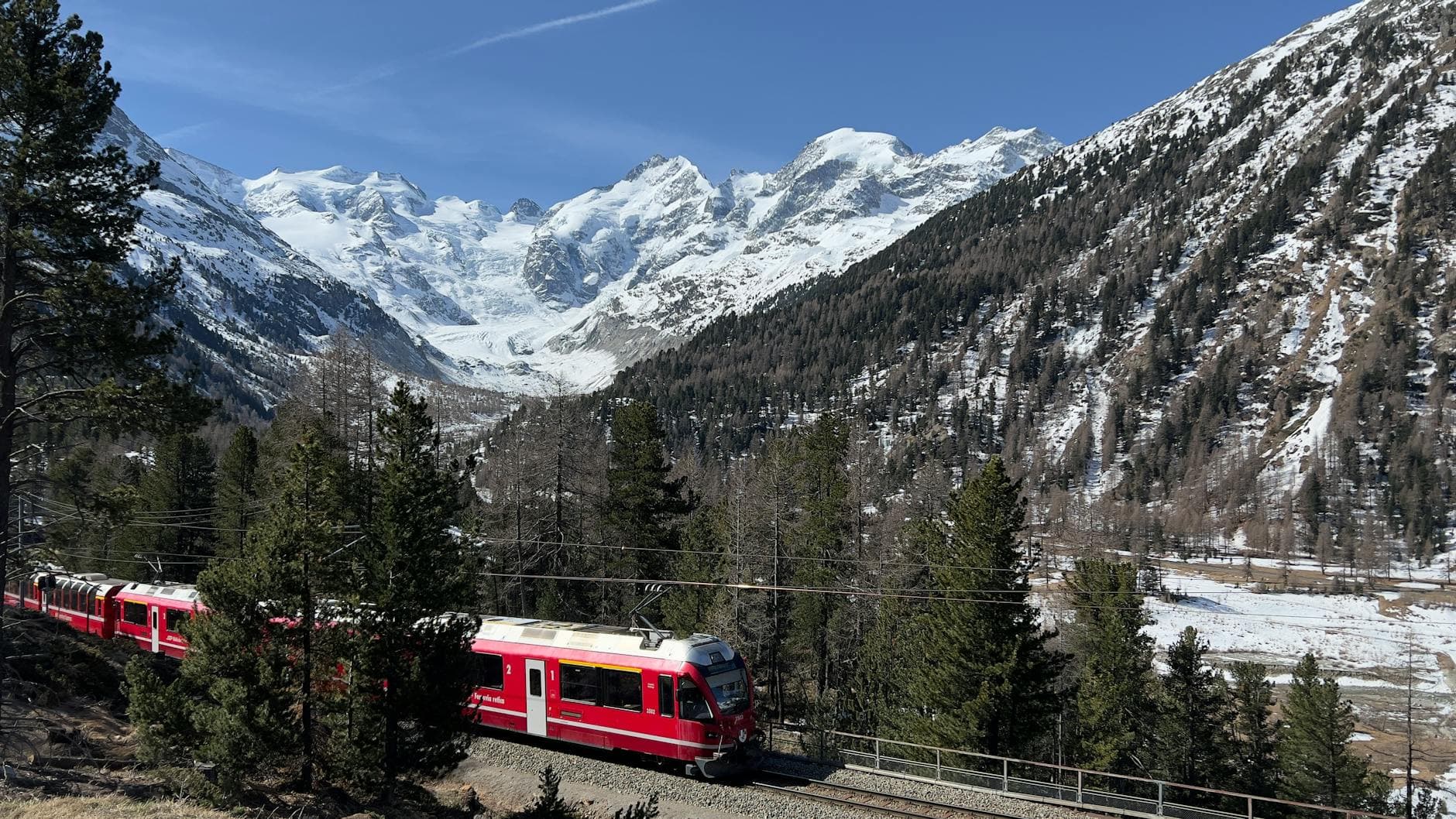 A red mountain train travels through an Alpine valley with snow-capped peaks, glaciers, and coniferous forests under clear blue sky. - Photo by Margis 233 on Pexels