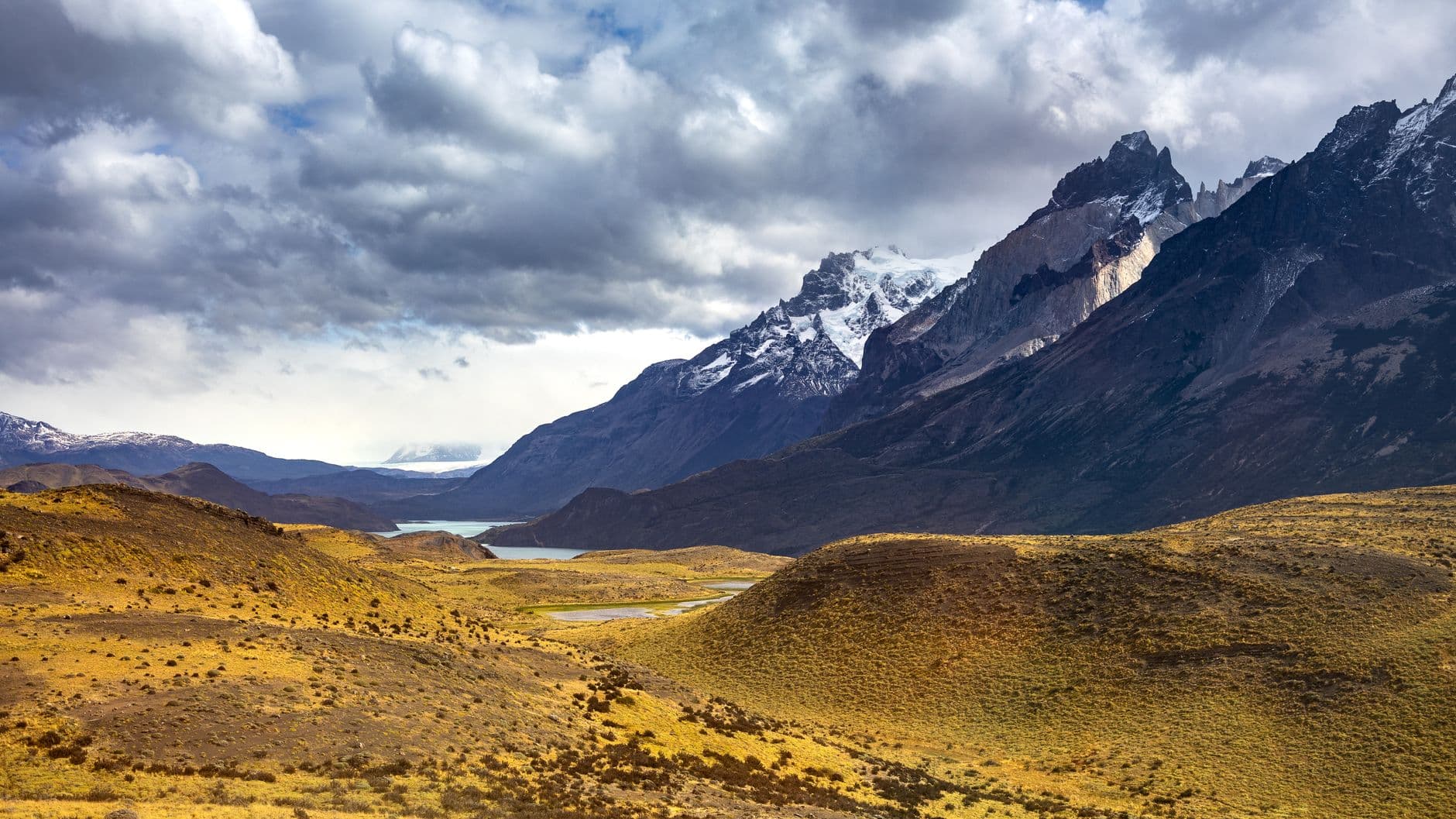 A panoramic view of the Torres del Paine landscape showing snow-capped granite peaks rising above a glacial valley with tundra vegetation, meltwater streams, and distant lakes under a cloudy Patagonian sky. - Photo by Marek Piwnicki on Pexels