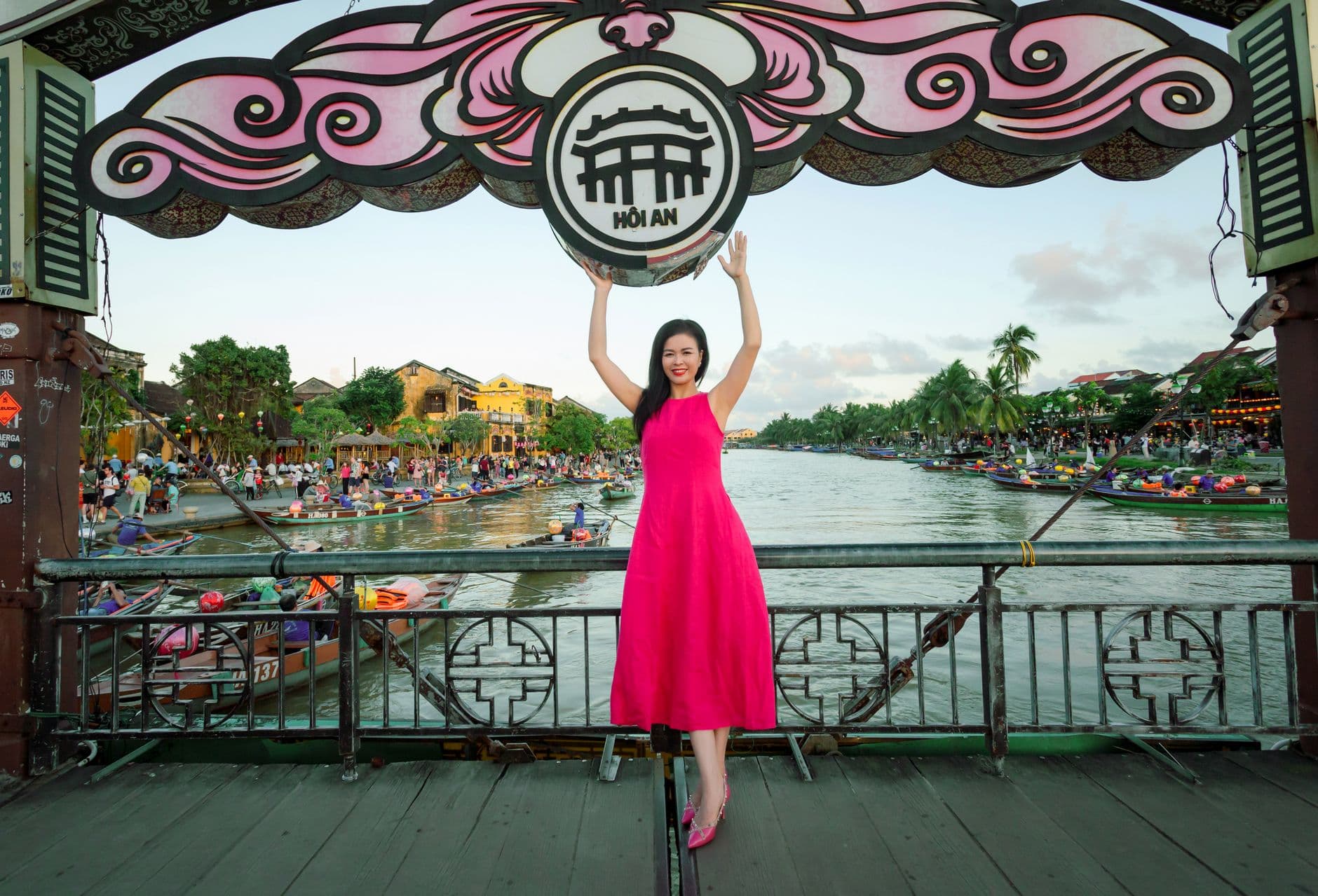 A woman in a bright pink dress poses on a wooden bridge over the Thu Bon River in Hoi An, Vietnam, holding up a decorative lantern sign, with the town's iconic yellow buildings and traditional boats visible in the background. - Photo by Võ Văn Tiến on Pexels