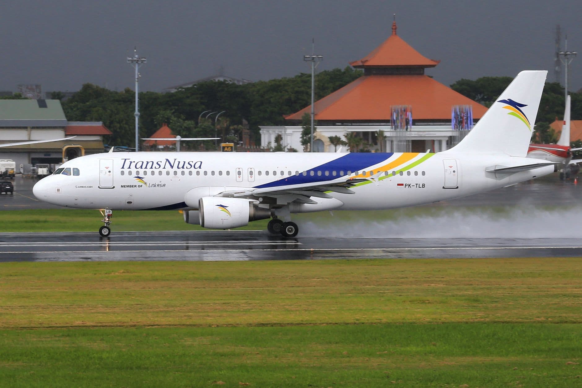 A TransNusa Airbus A320 aircraft with registration PK-TLB on a wet runway, with a distinctive orange-roofed building visible in the background. - Photo by Jeffry Surianto on Pexels