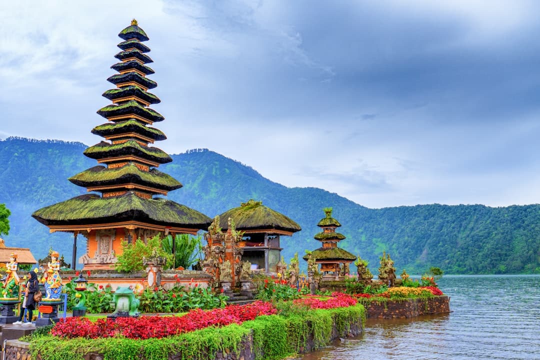 A striking Balinese Hindu temple complex set on a lake or waterfront with a prominent multi-tiered pagoda roof, ornate orange structures, flowering gardens, and forested volcanic mountains in the background. - Photo by Mahmud Ahsan on Unsplash
