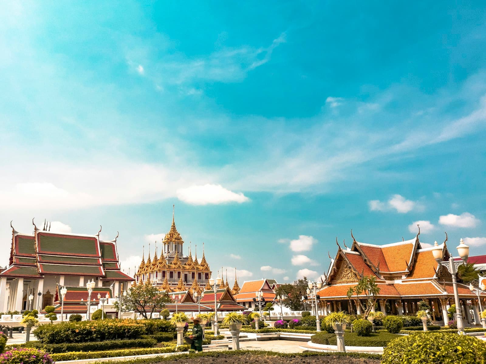 A complex of ornate Thai Buddhist temples with golden spires and traditional multi-tiered roofs with upturned eaves, surrounded by manicured gardens and landscaping under a clear blue sky. - Photo by Alejandro Cartagena 🇲🇽🏳🌈 on Unsplash