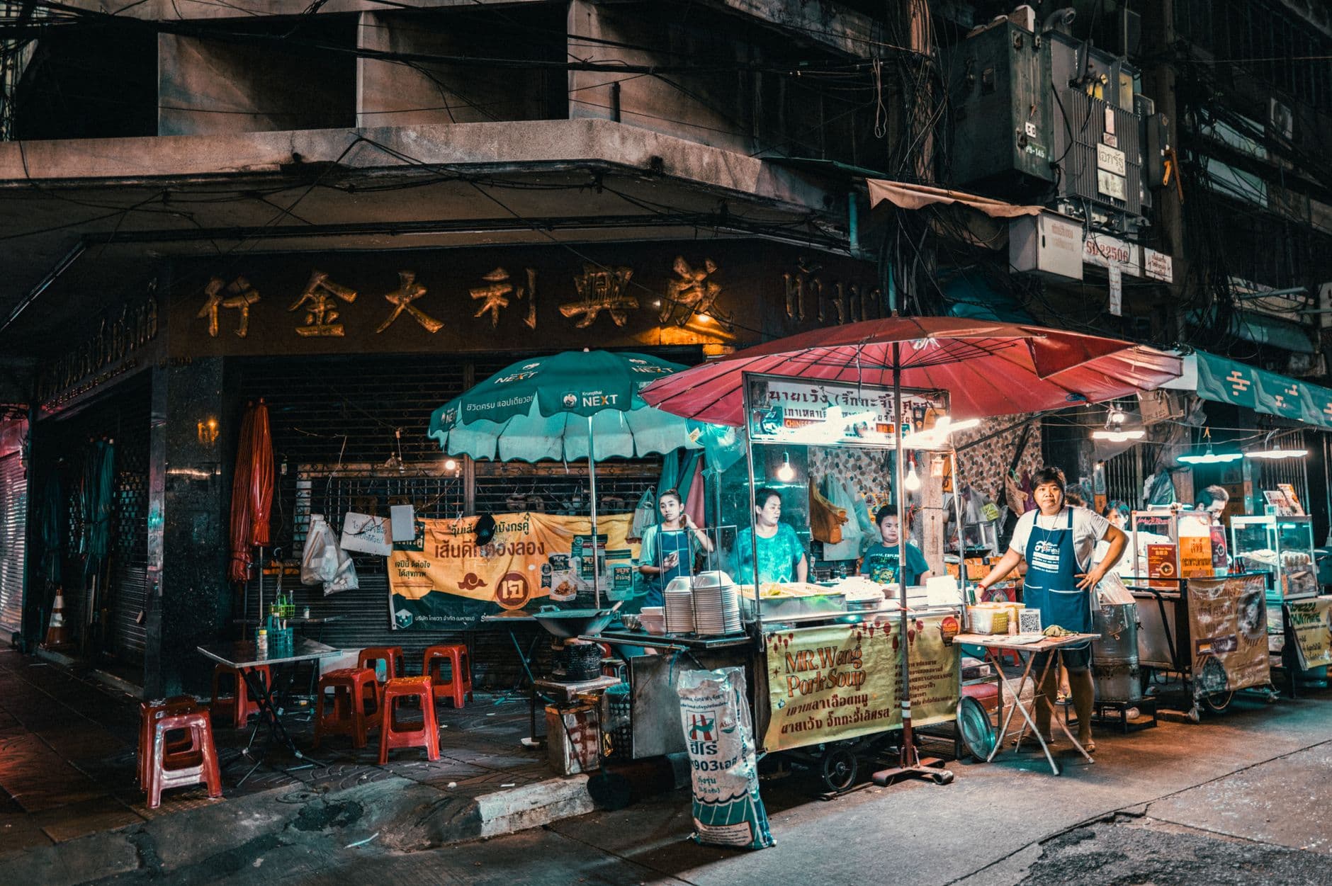 A street food vendor scene in Bangkok's Chinatown showing a small restaurant stall with teal umbrellas, red plastic stools, and Thai signage, representing the characteristic street food culture of Thailand's capital. - Photo by Tony Wu on Pexels