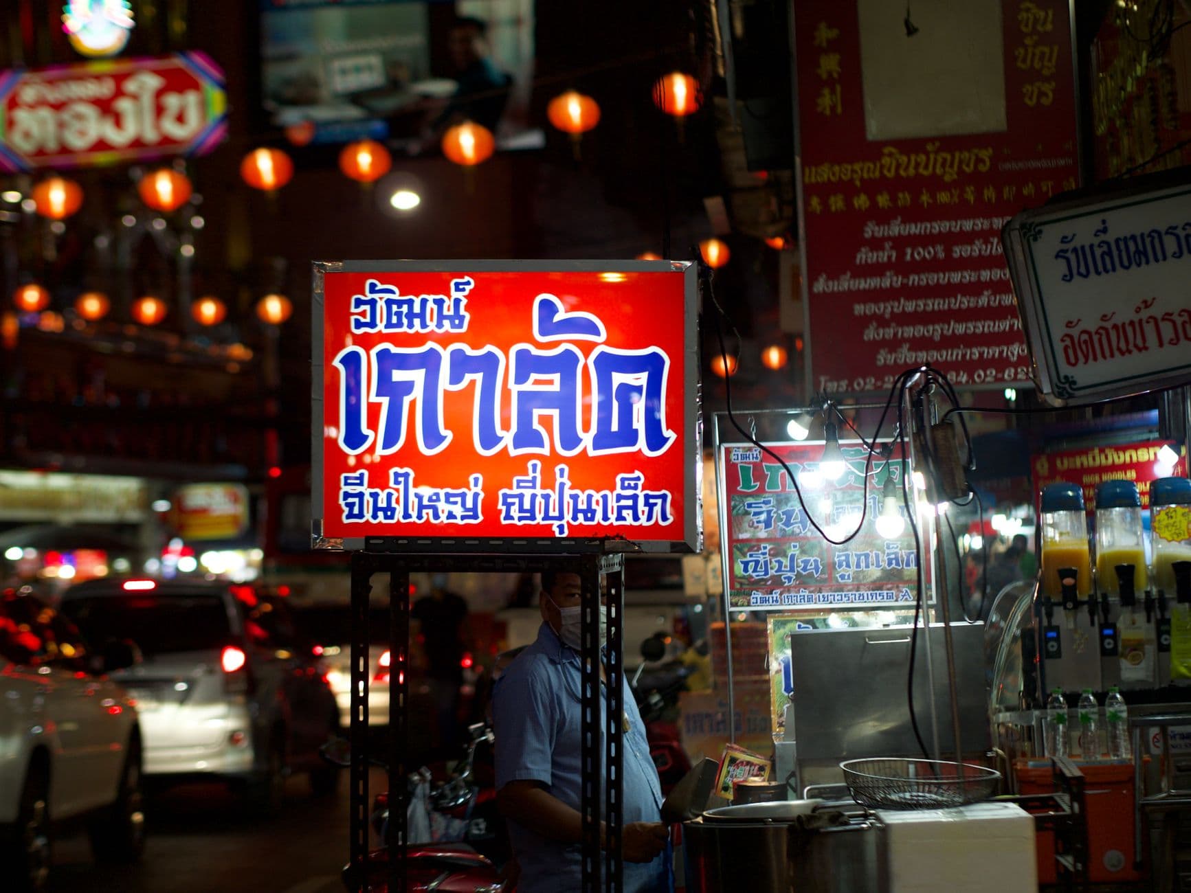 A night street food vendor stall in Bangkok displaying a prominent red and blue neon sign with Thai text, surrounded by orange lanterns, cooking equipment, and other storefronts with Thai signage. - Photo by Bobby Brown on Pexels