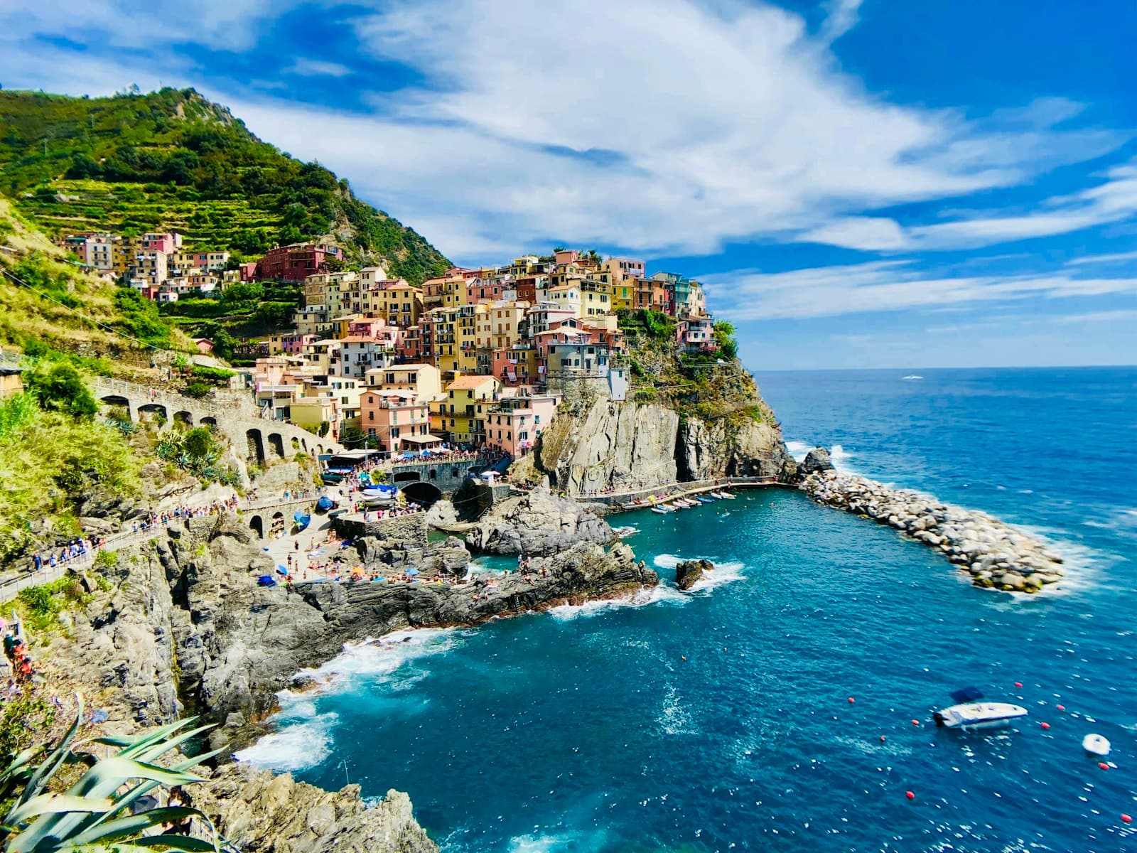 A picturesque cliffside village with colorful multi-story buildings cascading down a steep rocky promontory into a turquoise cove, with terraced vineyards above and the Mediterranean Sea below. - Photo by Mike Swigunski on Unsplash