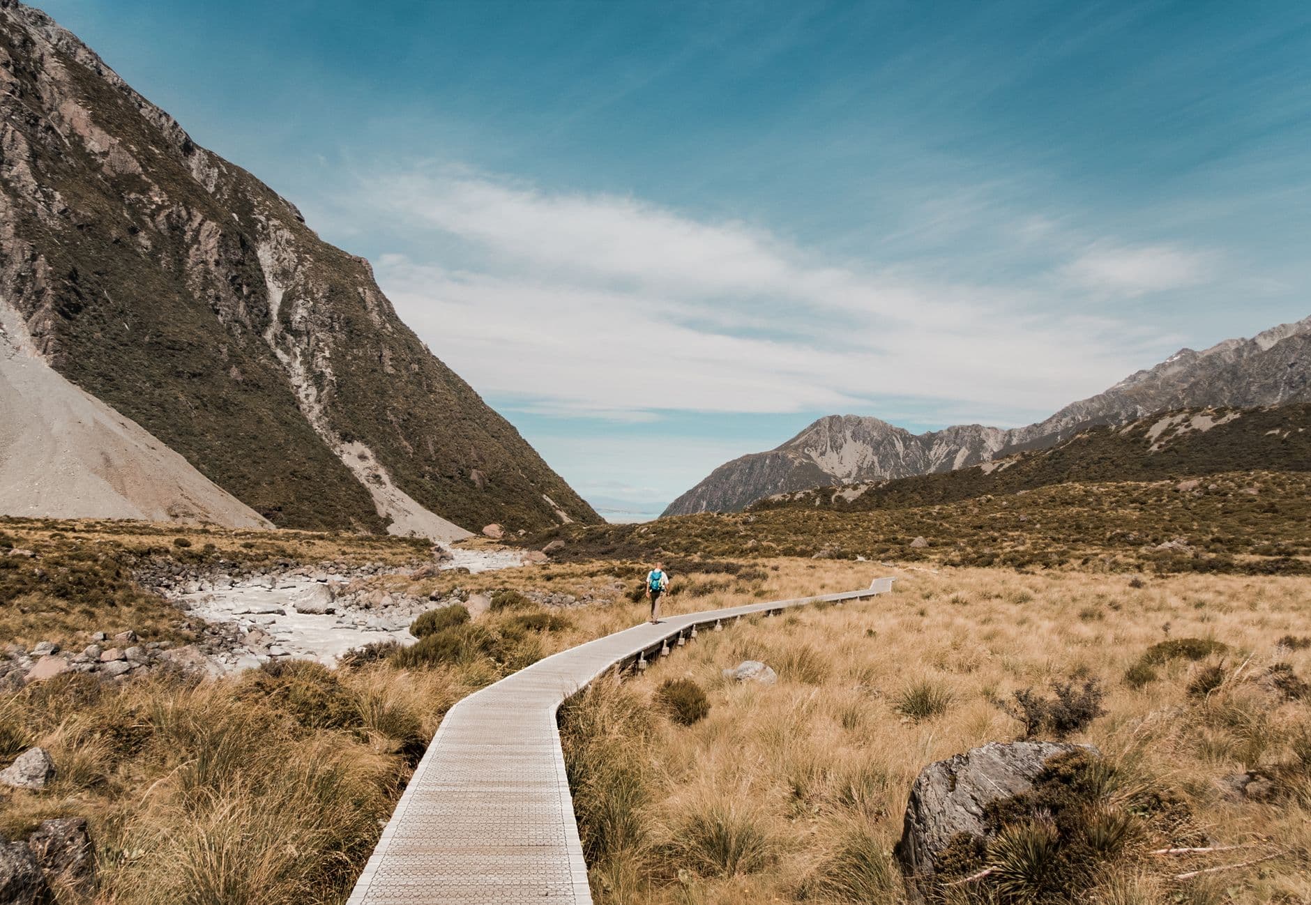 A hiker on a wooden boardwalk crosses alpine tussock grassland in a dramatic mountain valley with steep scree slopes, a glacial stream, and distinctive peaked mountains in the distance. - Photo by Tyler Lastovich on Pexels
