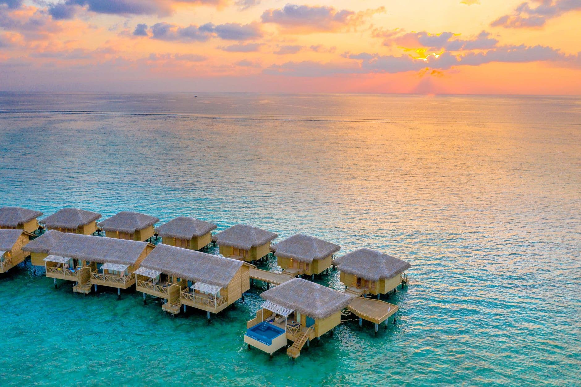 A row of overwater bungalows with thatched roofs at a luxury resort in the Maldives at sunset, showing the turquoise shallow water and typical island hospitality infrastructure. - Photo by Asad Photo Maldives on Pexels