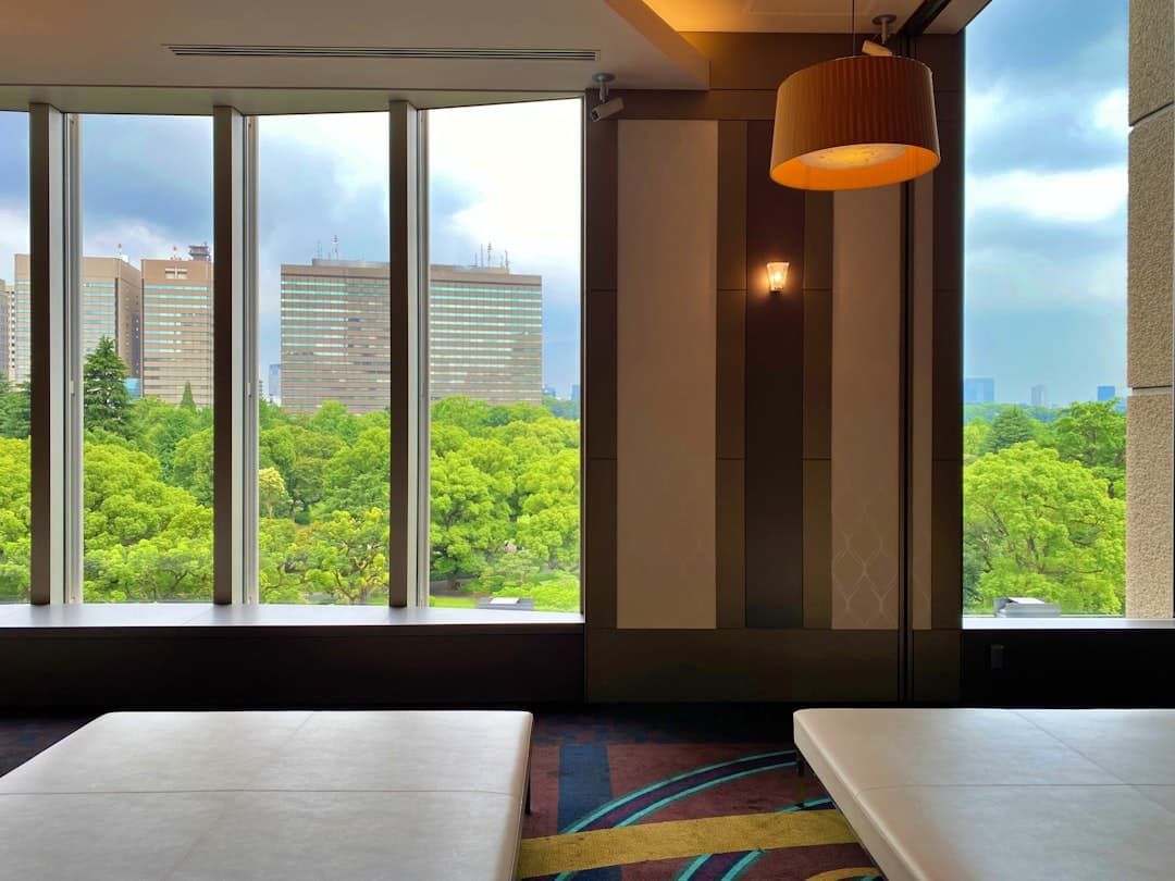 A modern hotel room or lounge interior with floor-to-ceiling windows overlooking a Tokyo cityscape with forested areas and contemporary office buildings. - Photo by Suzi Kim on Unsplash