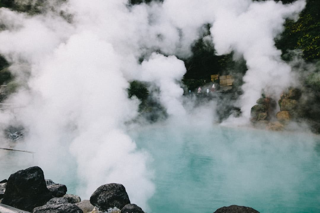 A geothermal hot spring pool with bright turquoise water shrouded in thick white steam and mineral vapor, with rocky outcrops in the foreground and Japanese-style wooden structures visible through the mist on the far shore. - Photo by Tayawee Supan on Unsplash