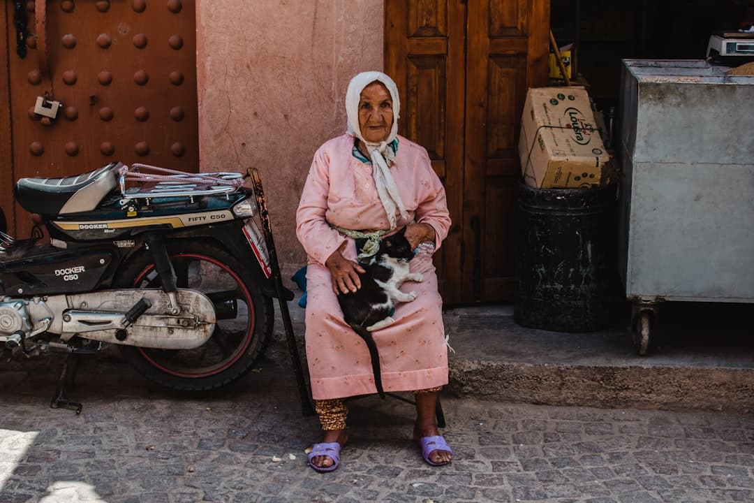 An elderly woman in traditional Moroccan dress sits on a street in a medina, holding a cat, surrounded by a motorcycle and household items against pink earthen walls. - Photo by Matthew Stephenson on Unsplash