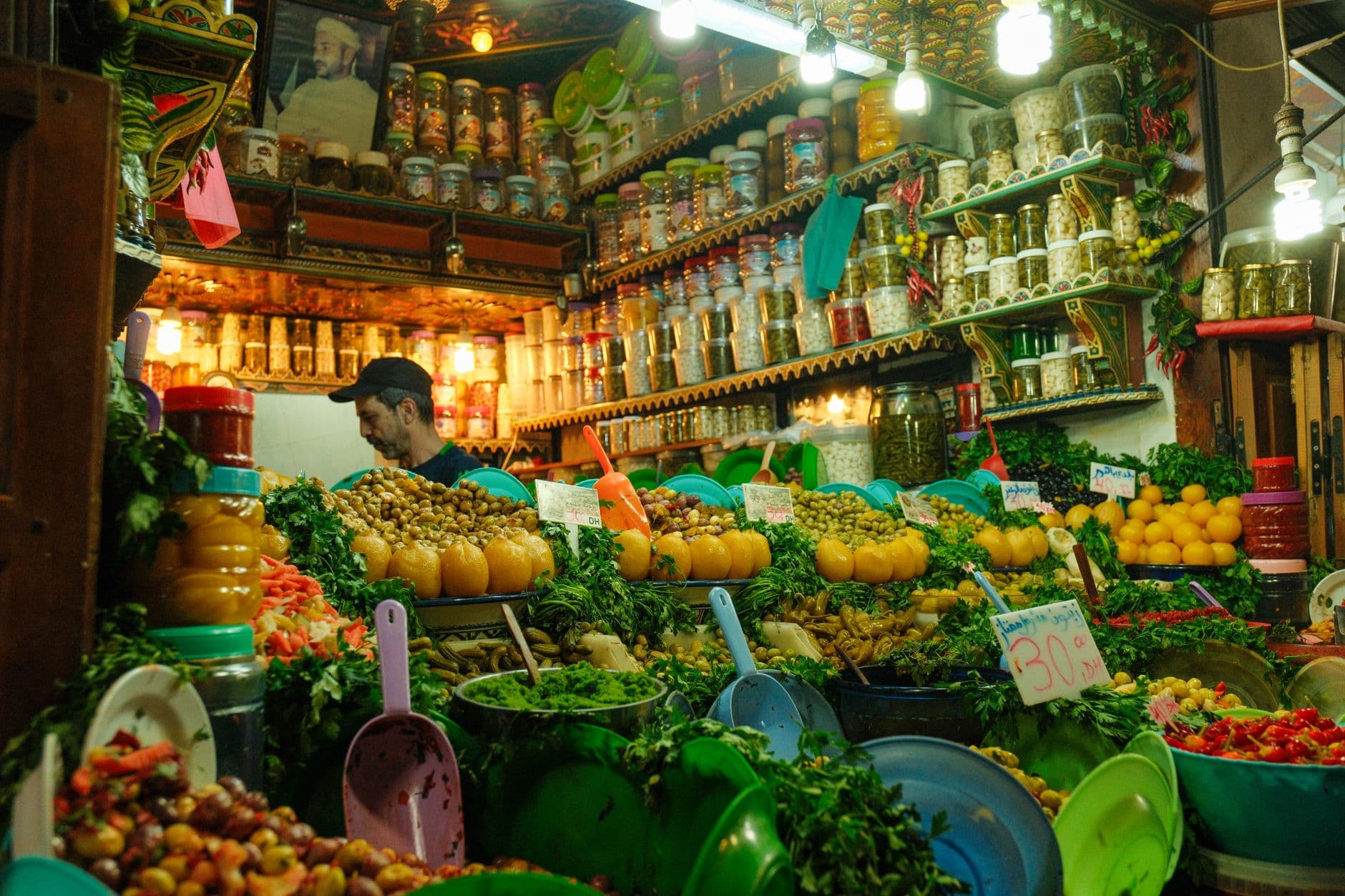A vibrant market stall in Fez's medina displaying preserved olives, lemons, fresh greens, and traditional Moroccan provisions with characteristic turquoise shelving and decorative ceiling elements. - Photo by Ramon Karolan on Pexels