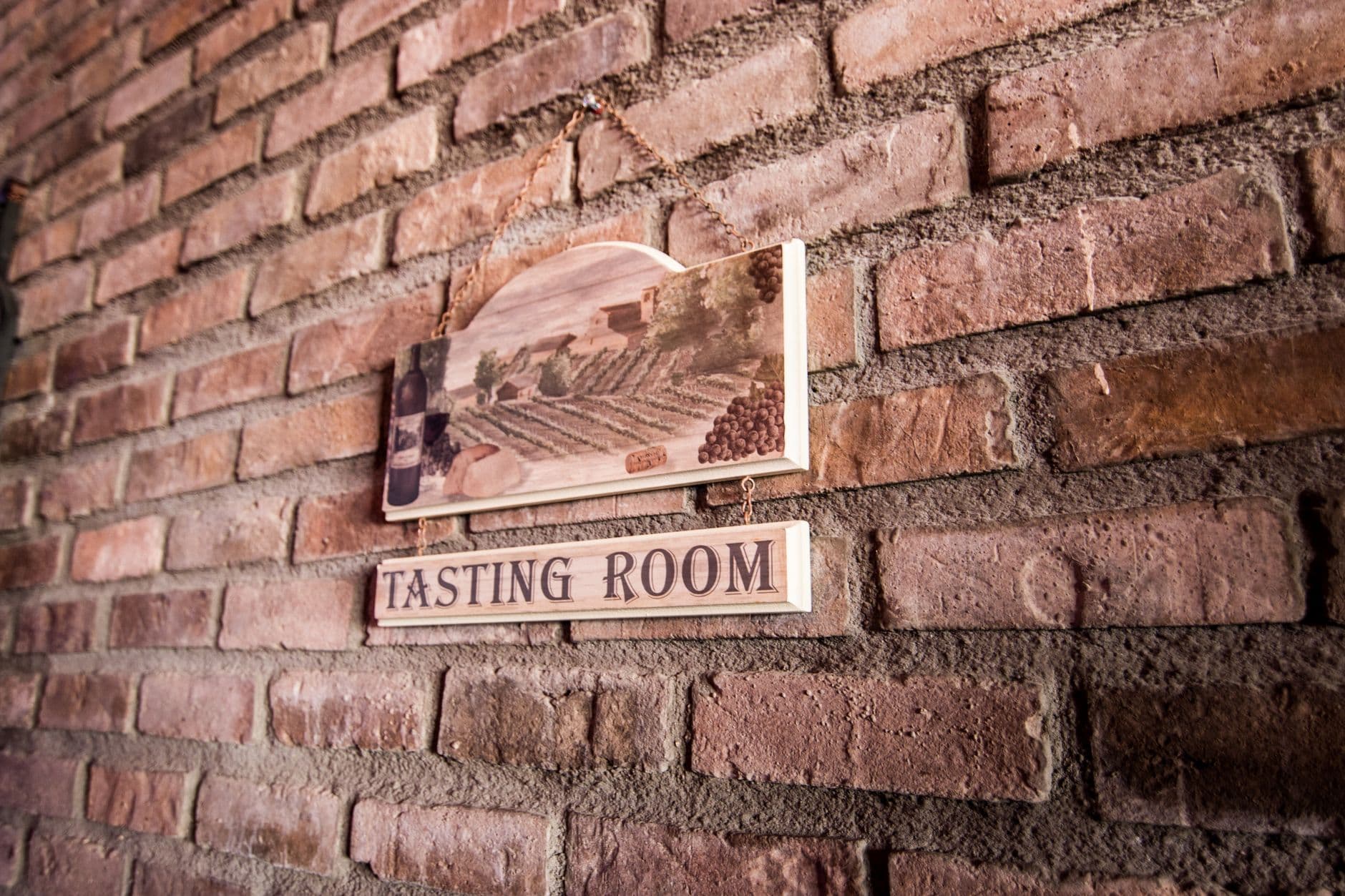 A wooden tasting room sign mounted on aged red brick wall, featuring an illustration of vineyard rows and grapes above the text. - Photo by Ash Craig on Pexels