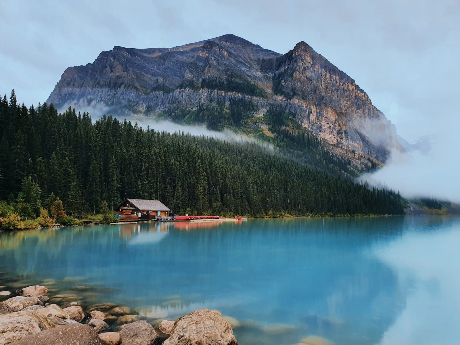 A pristine alpine lake with brilliant turquoise water reflects a dramatic rocky mountain peak shrouded in mist, with a red-roofed boathouse and canoes on the shore, surrounded by dense coniferous forest. - Photo by Fransiskus Filbert Mangundap on Unsplash