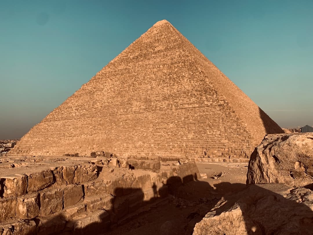 A direct frontal view of the Great Pyramid of Khufu at the Giza Plateau, photographed under clear skies with the limestone structure dominating the frame and desert terrain visible in the foreground. - Photo by ahmed ashraf on Unsplash