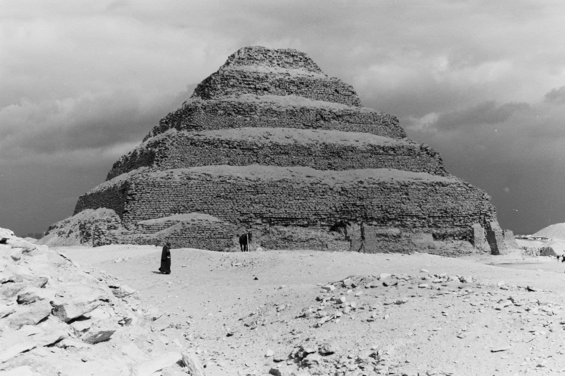 A black and white photograph of the Step Pyramid of Djoser at Saqqara, showing its iconic stepped limestone structure against an overcast sky, with desert floor in the foreground and small figures at its base for scale. - Photo by Dee Willi on Pexels