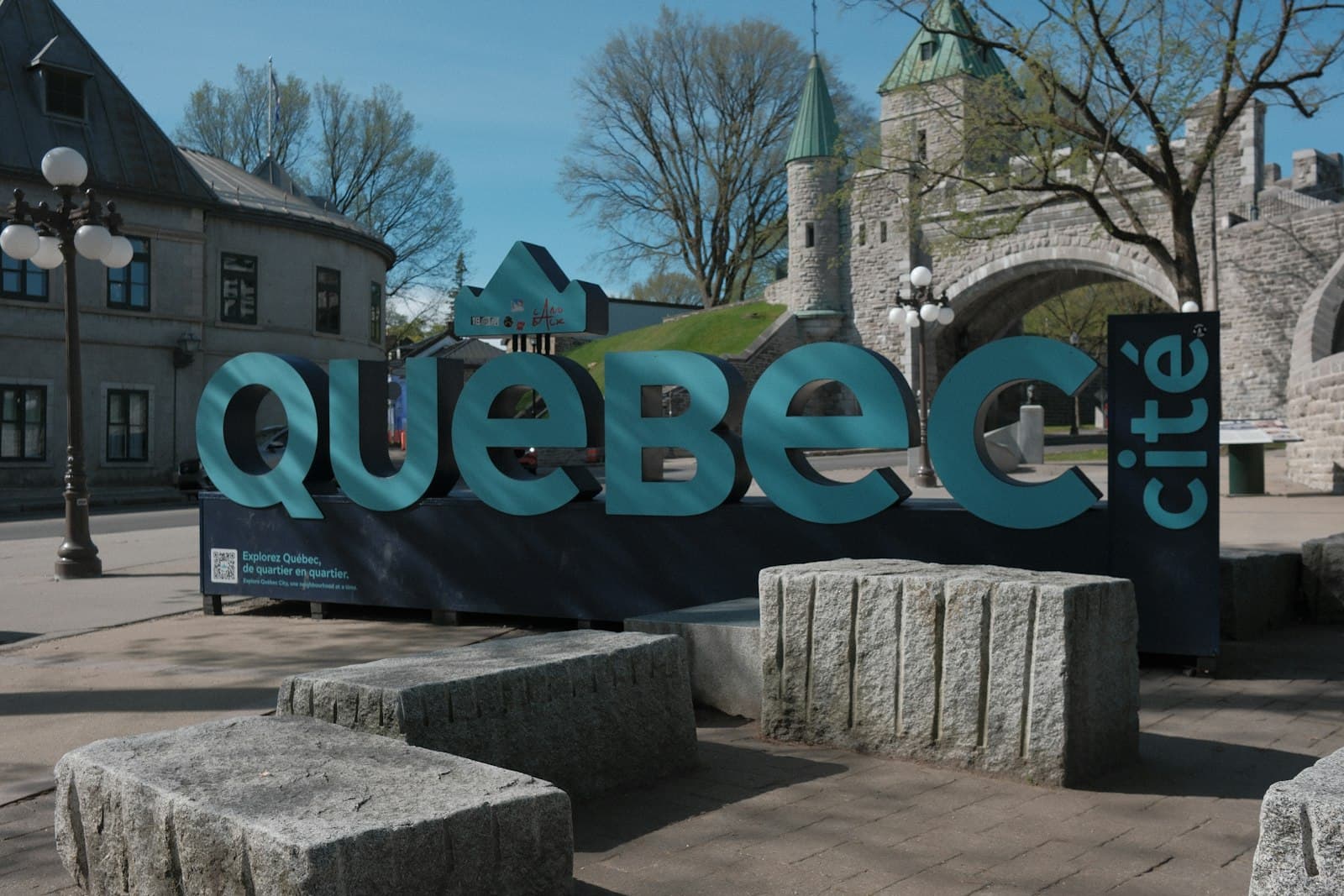 A large turquoise 'Quebec city' branded sign in a public plaza, with historic stone fortifications, a tower with green copper roof, and period architecture visible in the background. - Photo by Surinder Singh on Unsplash