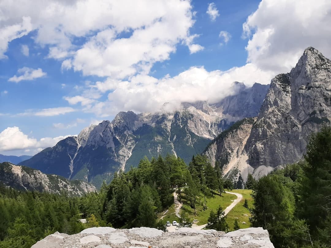 A dramatic alpine valley with steep limestone peaks, coniferous forest, a winding path or road, and a mountain stream, photographed under partly cloudy skies. - Photo by Mat Scott on Unsplash