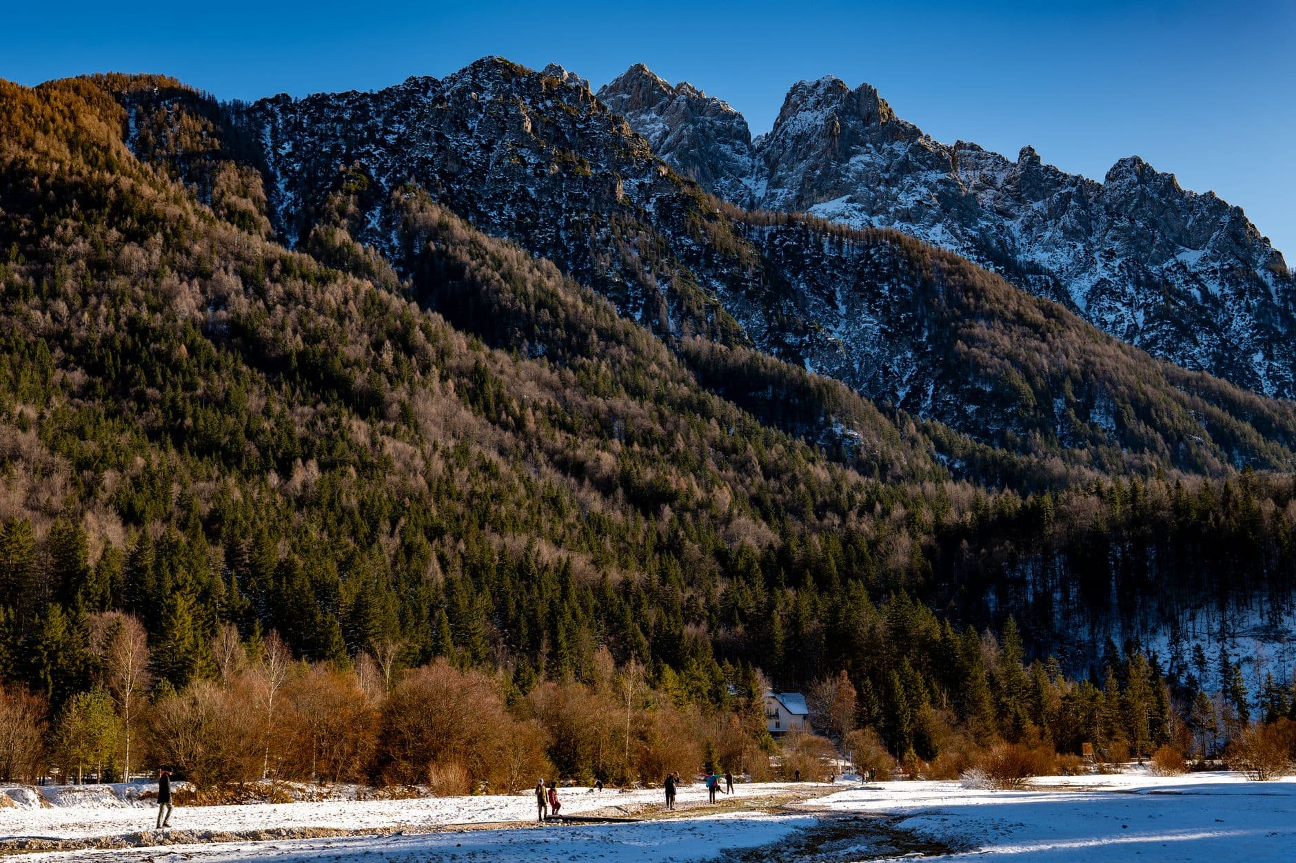 An autumn scene at an Alpine lake in the Julian Alps with forested foothills, snow-capped rocky peaks, and a few people on a snowy beach beside a partially frozen lake. - Photo by Vladimir Srajber on Pexels
