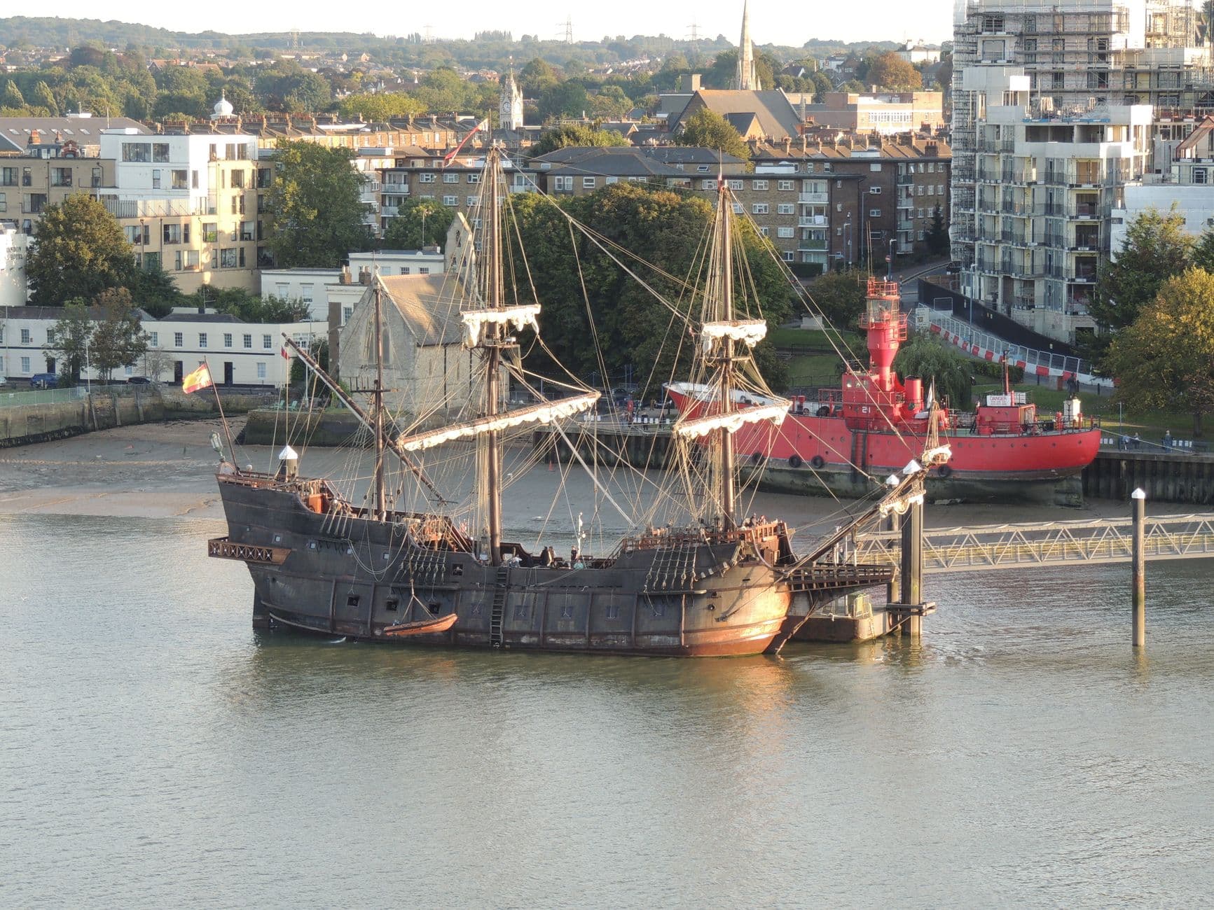 A historic tall ship with full rigging and wooden hull is moored on the River Thames in London, with contemporary apartment buildings, a red working vessel, and church spires visible in the background. - Photo by M. Yates on Pexels