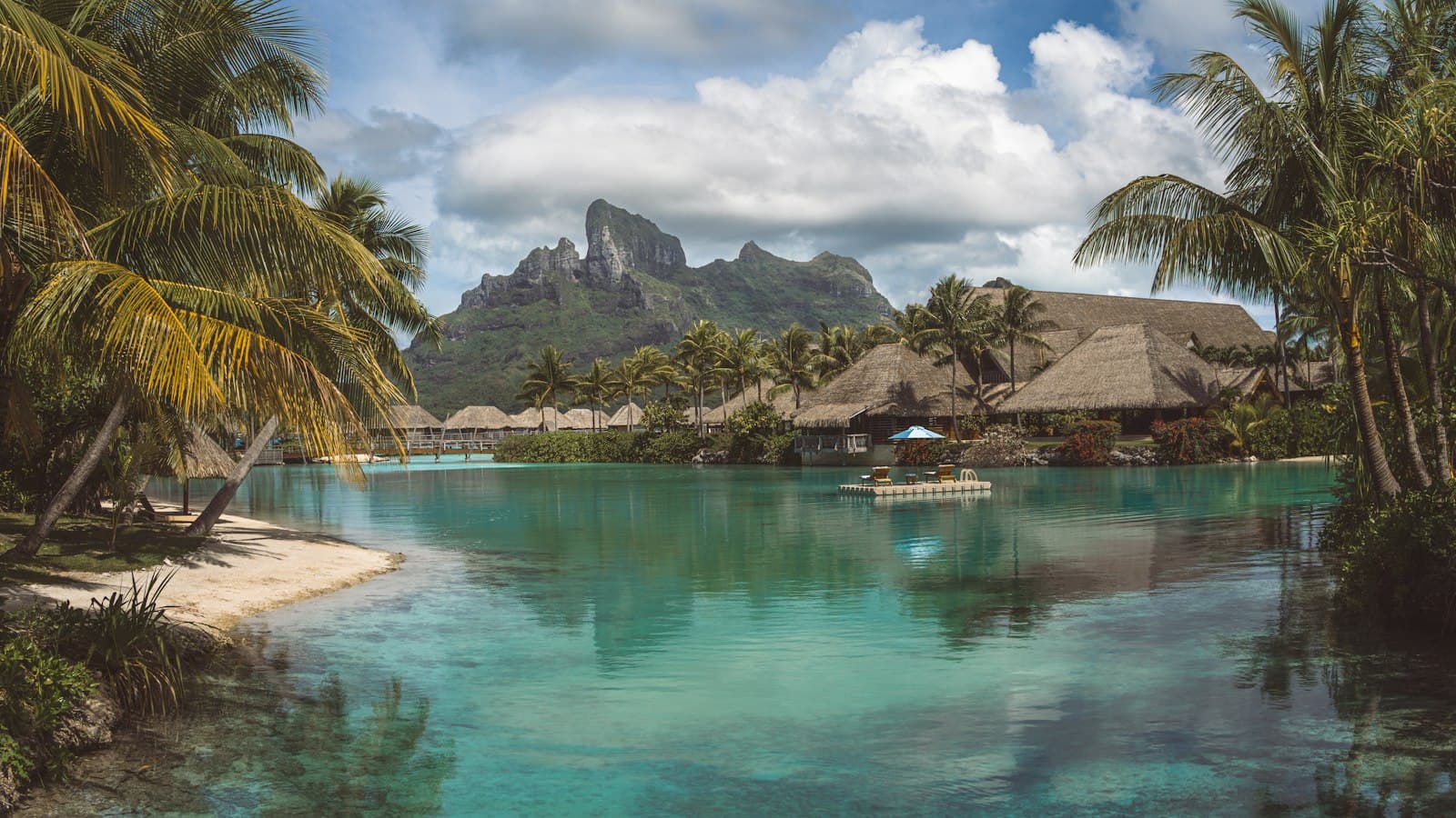 A luxury overwater resort in a turquoise lagoon with thatched-roof bungalows, coconut palms, and Bora Bora's distinctive peaked mountain backdrop. - Photo by john ko on Unsplash