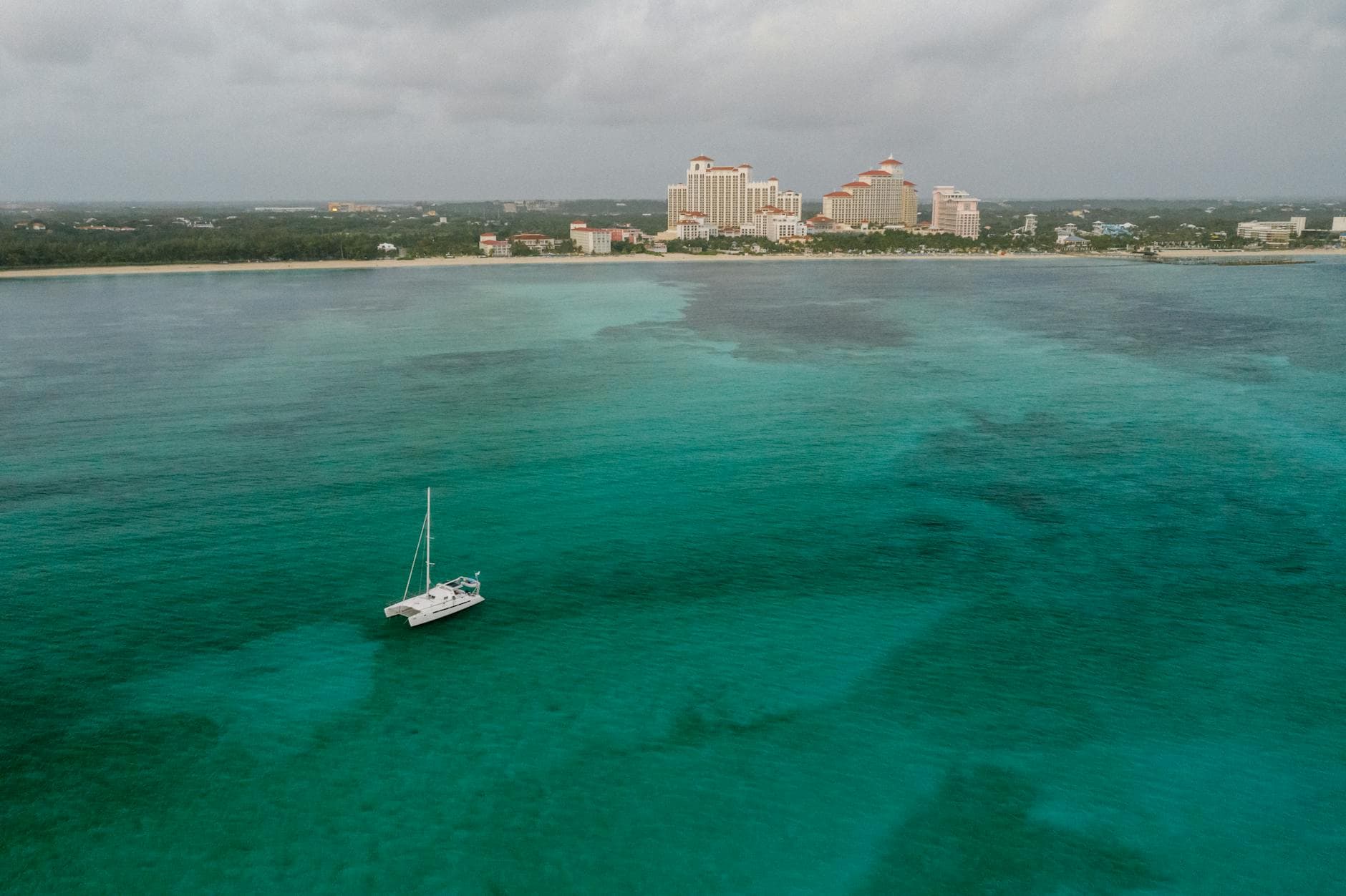 A sailboat anchors in clear turquoise water off a sandy beach, with a large resort development featuring distinctive copper-roofed buildings visible along the shoreline. - Photo by Mikhail Nilov on Pexels