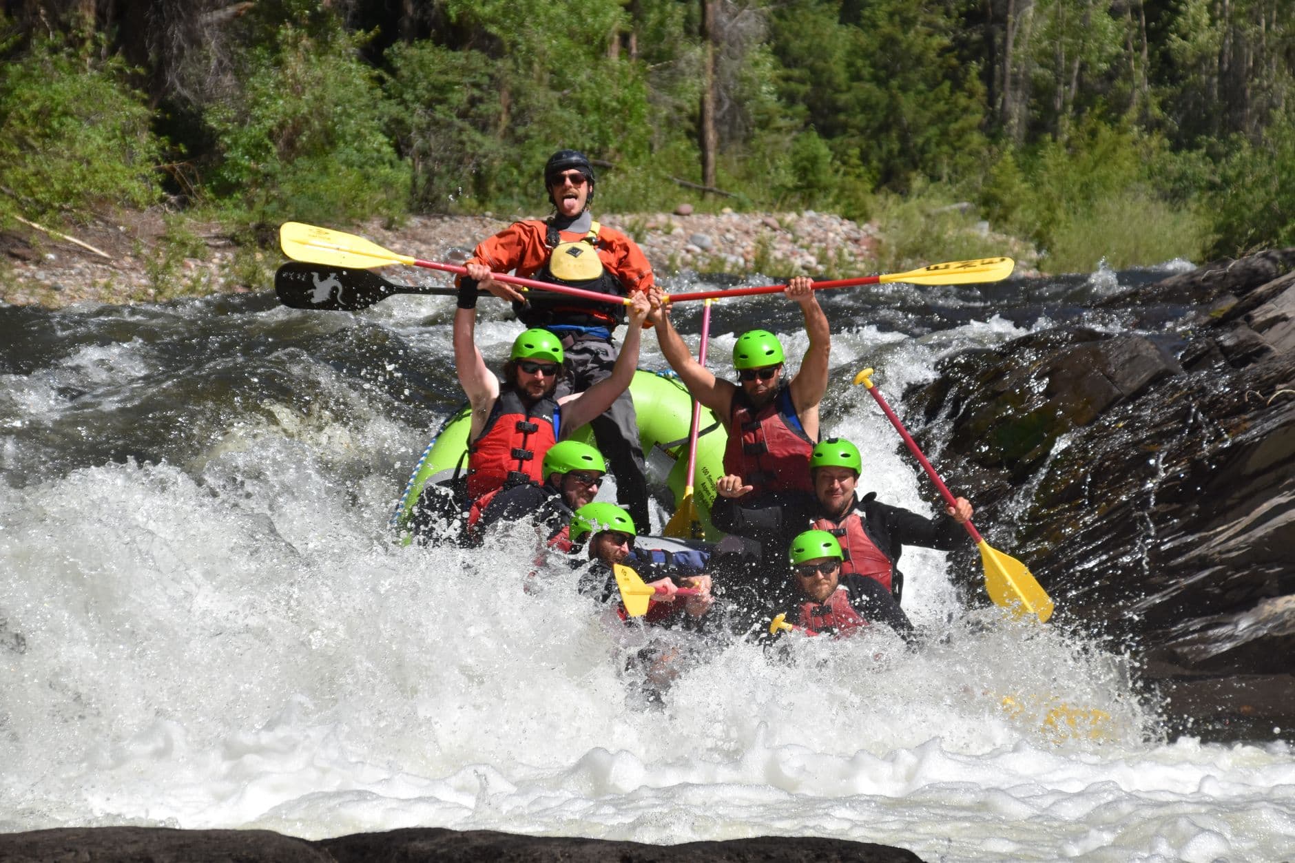 A commercial whitewater rafting group navigates powerful rapids in a large raft, with multiple participants wearing bright green helmets and red/orange PFDs in heavy white water with coniferous forest visible on steep canyon walls. - Photo by David Hadley on Pexels