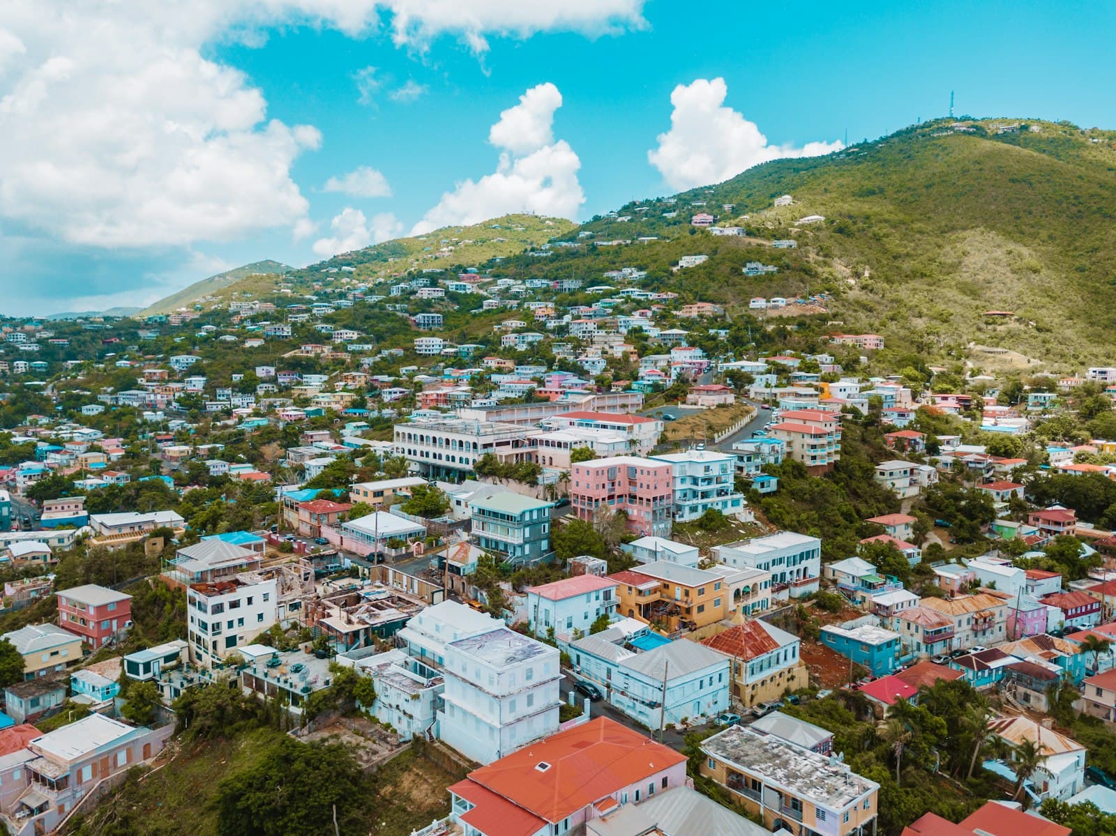Aerial view of a densely built hillside Caribbean town with colorful residential and commercial buildings scattered across steep terrain, with green hills rising behind. - Photo by Andy Feliciotti on Unsplash