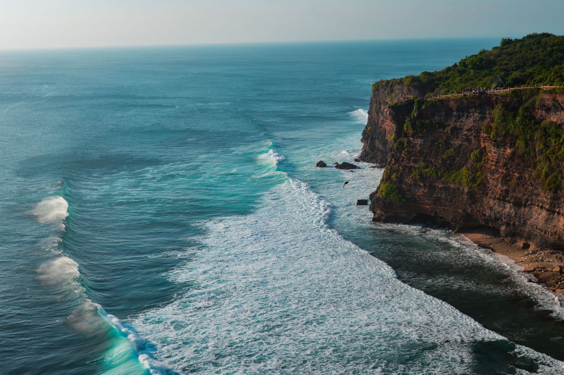 A dramatic coastal clifftop view showing steep volcanic cliffs covered in lush green vegetation overlooking turquoise ocean waters with white-capped waves, featuring a wooden safety railing along the cliff edge. - Photo by M-DESIGNZ LLC on Pexels