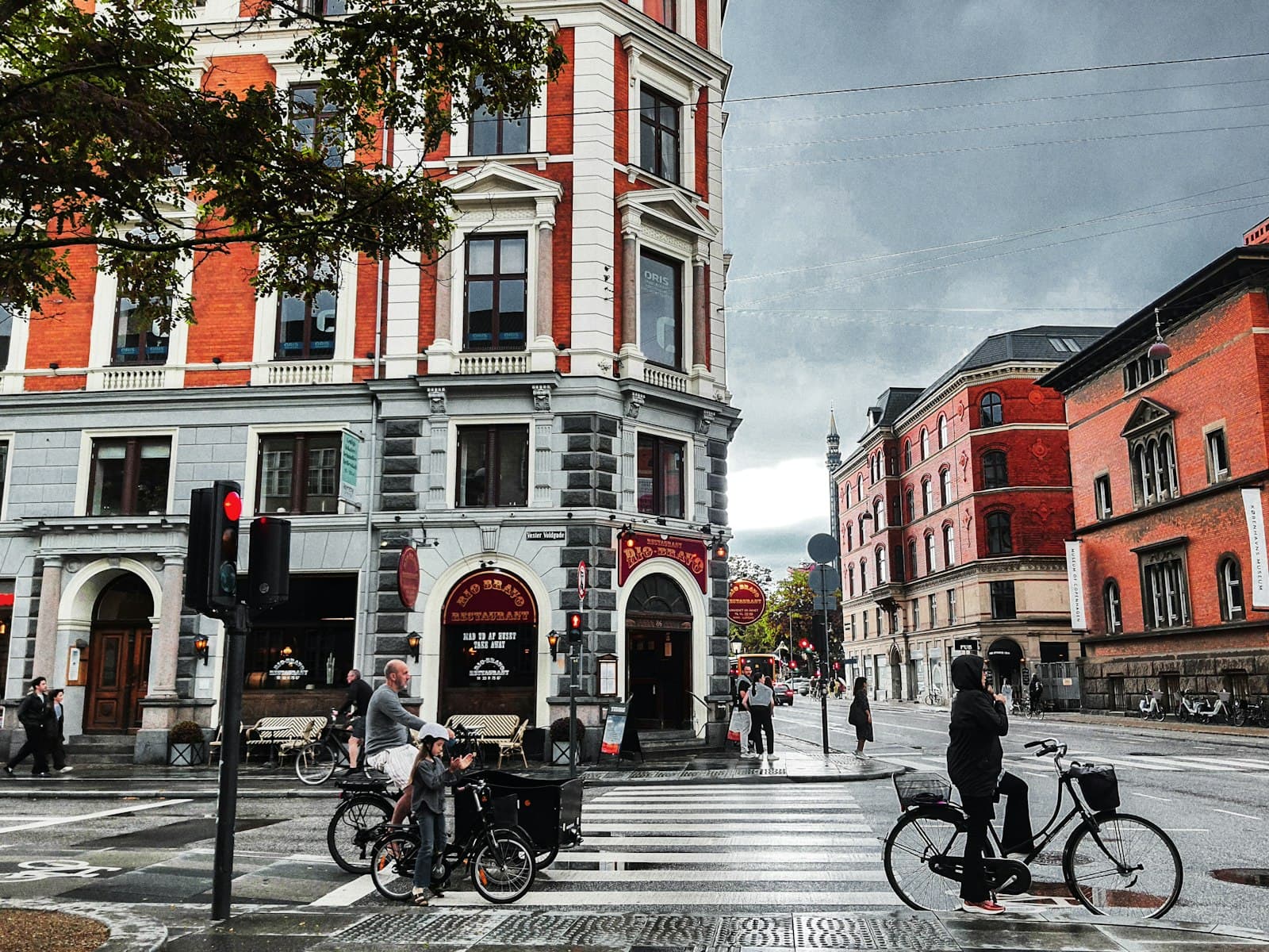 A wet Copenhagen street scene showing red-brick Victorian buildings, pedestrians, cyclists, and a busy urban plaza with characteristic Danish cycling culture and architecture. - Photo by flickch on Unsplash