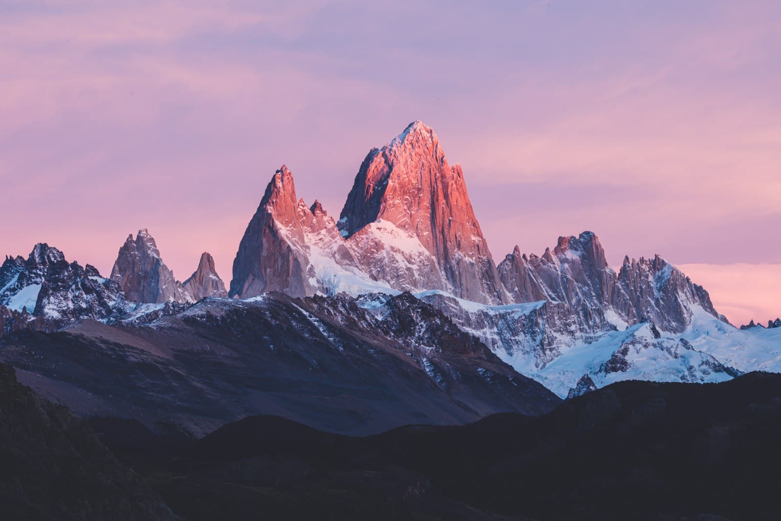 Dawn or dusk alpenglow illuminates the distinctive granite spires of Mount Fitz Roy and surrounding peaks in Patagonia, with snow-covered slopes and dark forested foothills in the foreground. - Photo by Andrew Svk on Unsplash