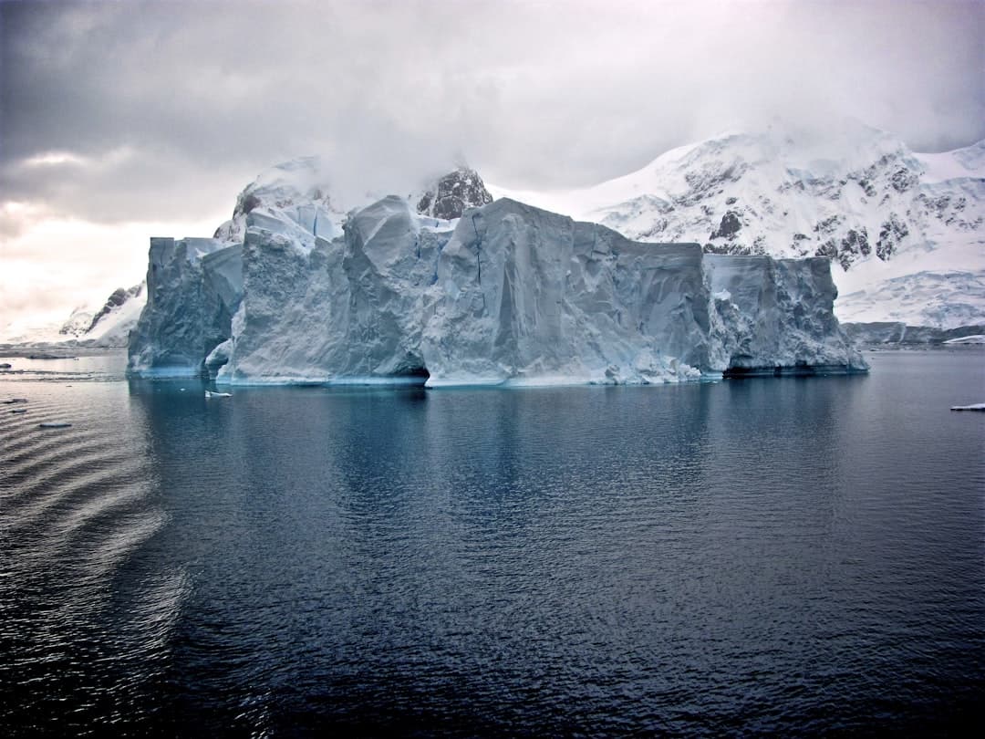 A large sculptured iceberg with cavernous formations floats in dark Antarctic waters with snow-covered peaks visible in the background. - Photo by Jay Ruzesky on Unsplash