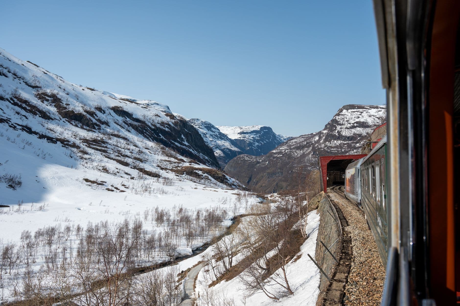 A red Norwegian passenger train travels through a snow-covered mountain valley with steep, forested slopes and a river, characteristic of the Flåm Railway scenic route in Norway. - Photo by Nils R on Pexels