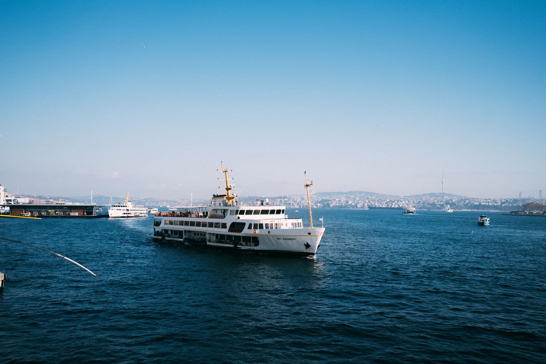 A white passenger ferry boat anchored in a strait with a densely built hillside city visible across the water under clear blue skies. - Photo by Sami TÜRK on Pexels