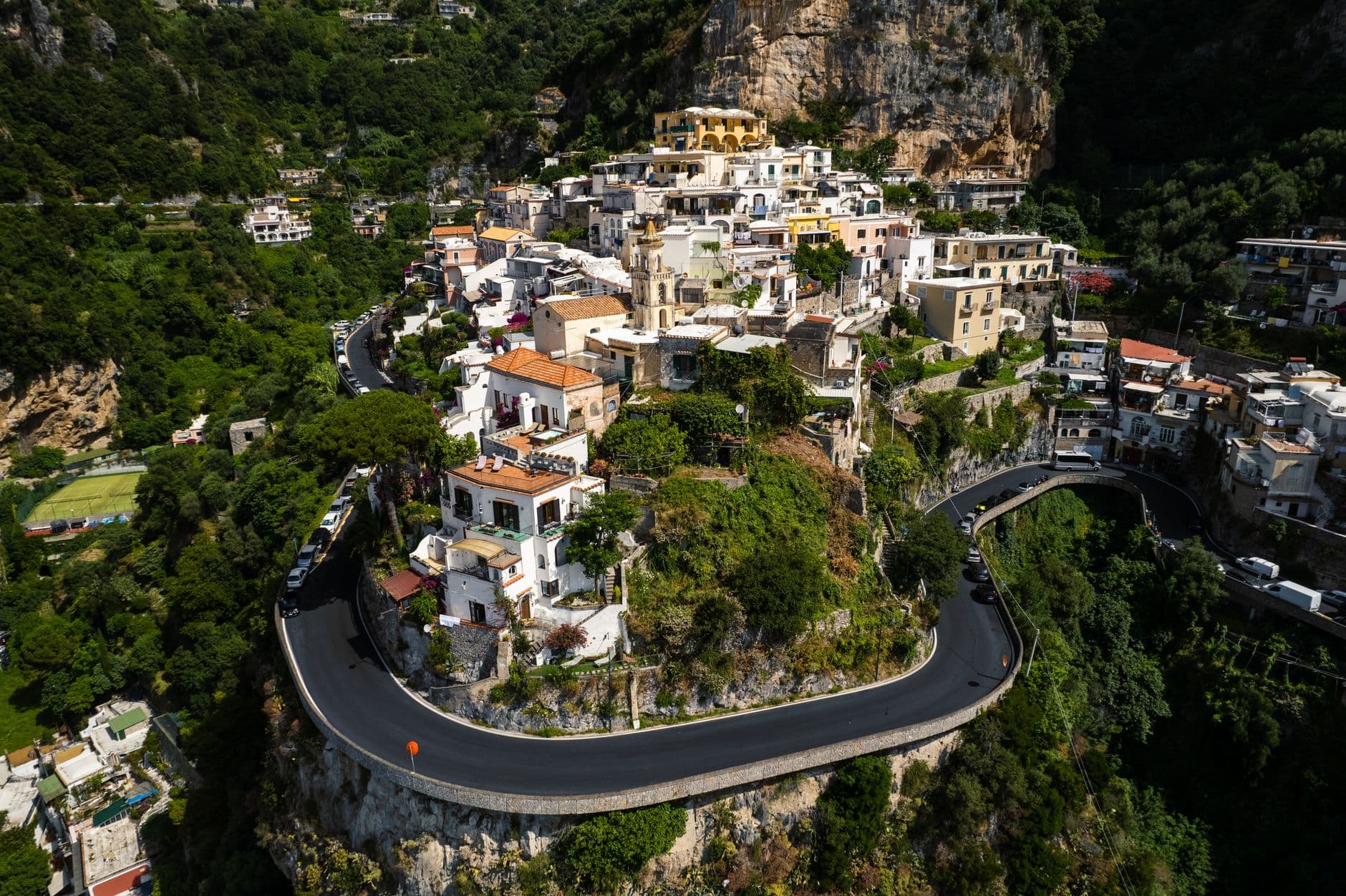 Aerial view of Positano village clinging to dramatic limestone cliffs on the Amalfi Coast, showing whitewashed and pastel-colored buildings cascading down the hillside, with a serpentine coastal road carved into the cliff face and lush green vegetation covering the surrounding terrain. - Photo by Matteo Parisi on Pexels