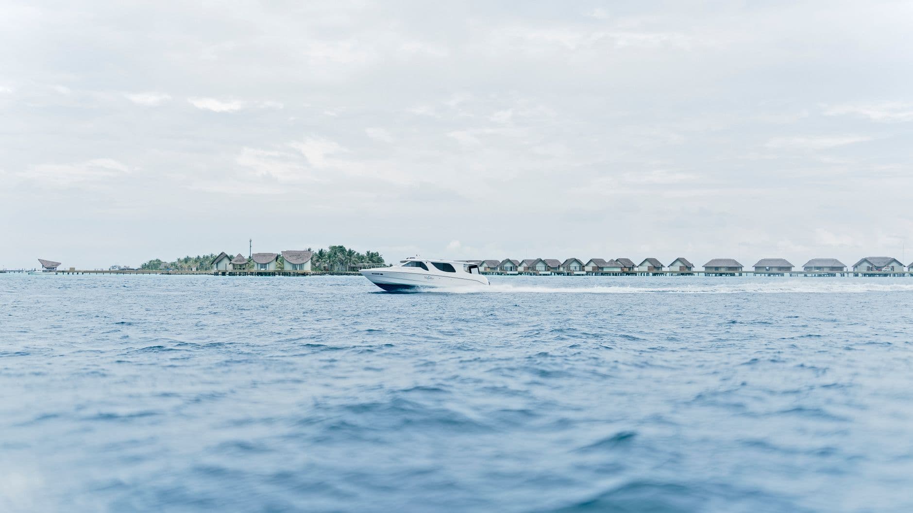A speedboat cruises past a row of over-water bungalows on a tropical atoll, with a resort island visible in the background. - Photo by Musaddek Sayek on Pexels