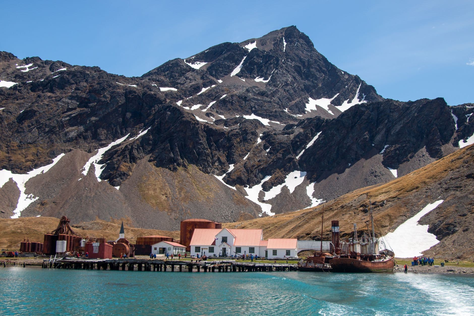 The historic whaling station at Grytviken, South Georgia, featuring the distinctive pink-roofed church, abandoned industrial structures, and a beached/moored rusted vessel, set against snow-dusted mountains and turquoise waters. - Photo by ArcticDesire.com Polarreisen on Pexels