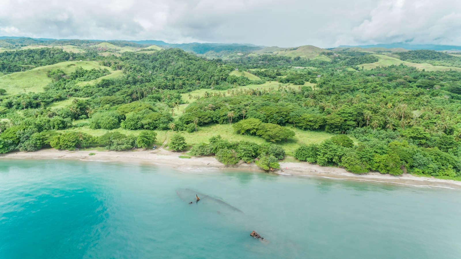 Aerial view of a pristine tropical coastline with dense rainforest, a turquoise lagoon containing visible submerged objects (possibly WWII wreckage), and sandy beaches backing onto lush green hills. - Photo by Gilly Tanabose on Unsplash