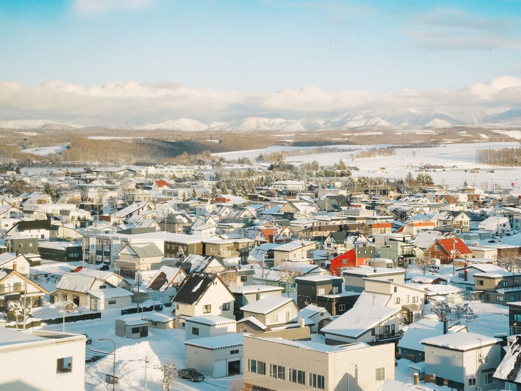 A snow-covered residential town in winter with snow-laden roofs, a mix of residential buildings in muted colors, surrounded by frozen agricultural fields and forested areas, with snow-capped mountains visible in the distant background. - Photo by Maxine Xin on Pexels