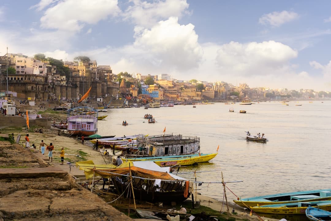 A view of Varanasi's waterfront along the Ganges River showing traditional colorful wooden boats, bathing ghats, pilgrims on the steps, and the characteristic densely-packed architecture of the old city rising up the hillside. - Photo by Kishor Kumar C K on Unsplash
